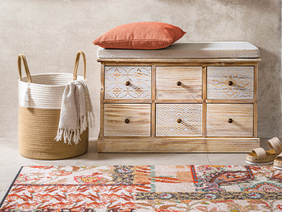 Meuble en bois avec motifs gravés, panier en corde avec plaid blanc, coussin orange et tapis coloré.