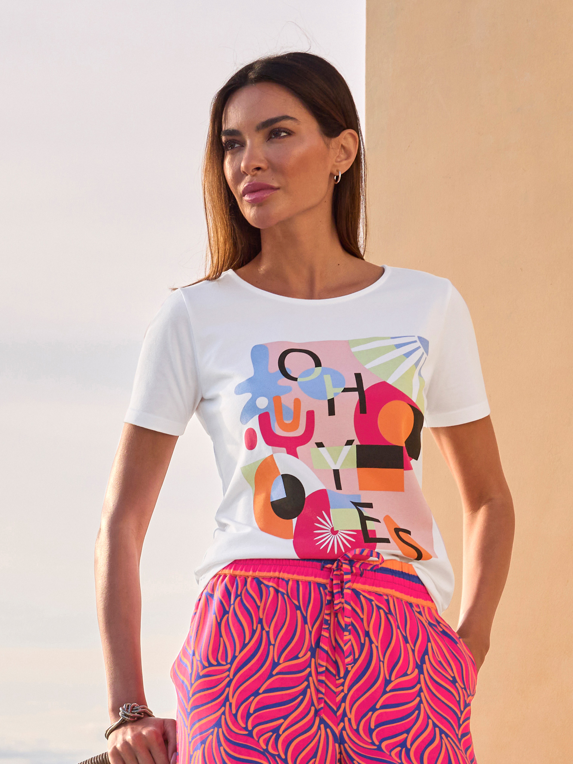 Un modèle porte un t-shirt blanc avec un imprimé abstrait coloré, associé à un pantalon rose-rouge à motif.