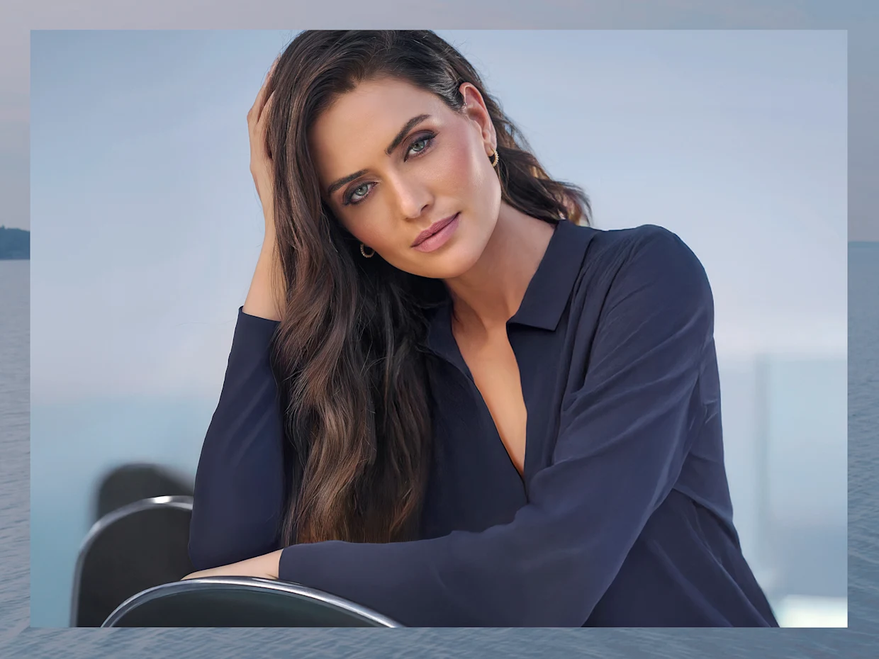 Ein Model mit langen braunen Haaren trägt ein dunkelblaues Oberteil und eine goldene Halskette, sitzt vor einem Wasserhintergrund. Ein Model mit langen braunen Haaren trägt ein dunkelblaues Oberteil und eine goldene Halskette, sitzt vor einem Wasserhintergrund.