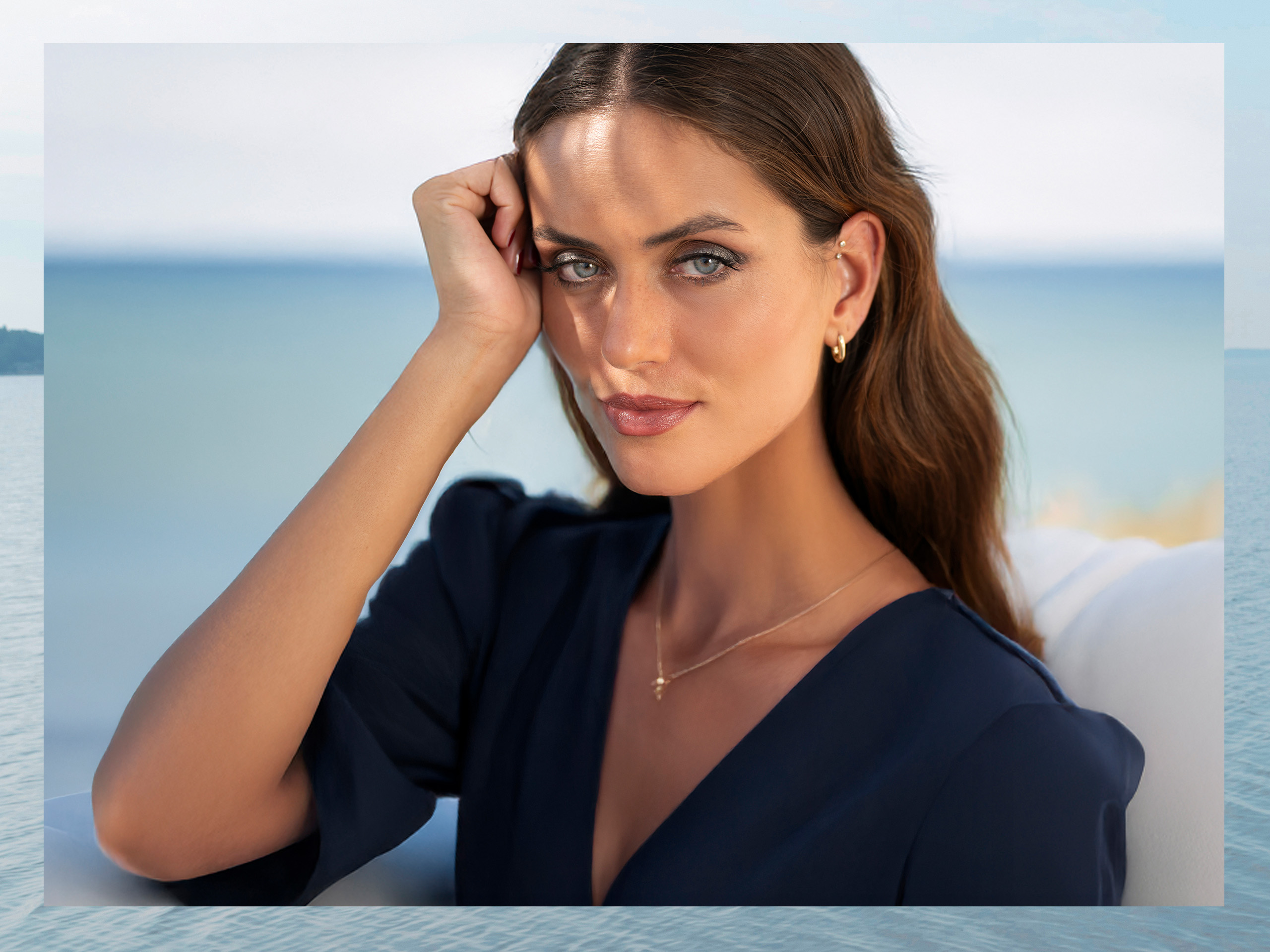 Een model met lang bruin haar draagt een donkerblauw bovenstuk en een gouden ketting, zittend voor een waterachtergrond.