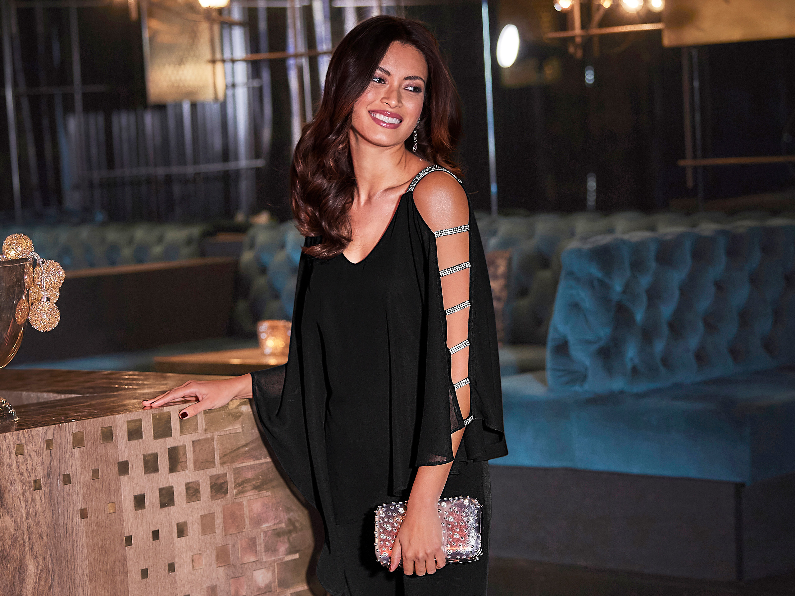 Een model draagt een zwarte jumpsuit met open mouwen die met zilveren bandjes aan elkaar zijn verbonden, en houdt een glinsterende clutch in haar hand.