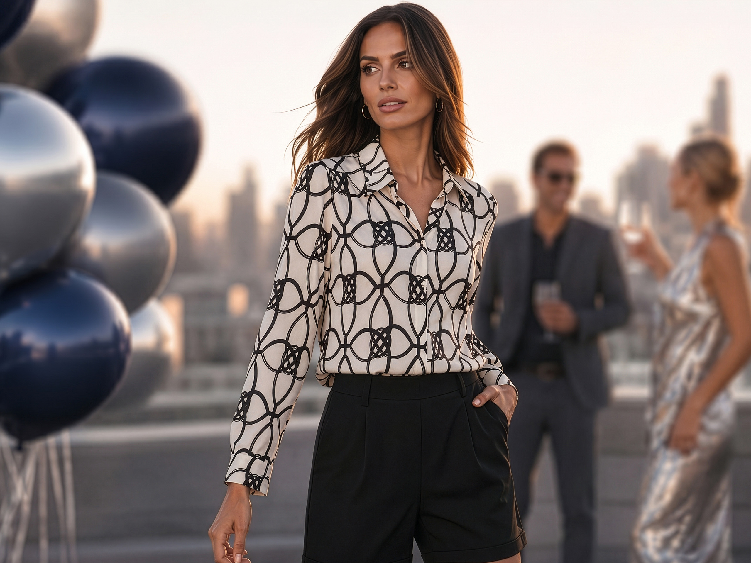 Un mannequin porte une blouse blanche à motif chaîne noire et un short noir, debout sur une terrasse sur le toit, en arrière-plan deux personnes floues et des ballons.