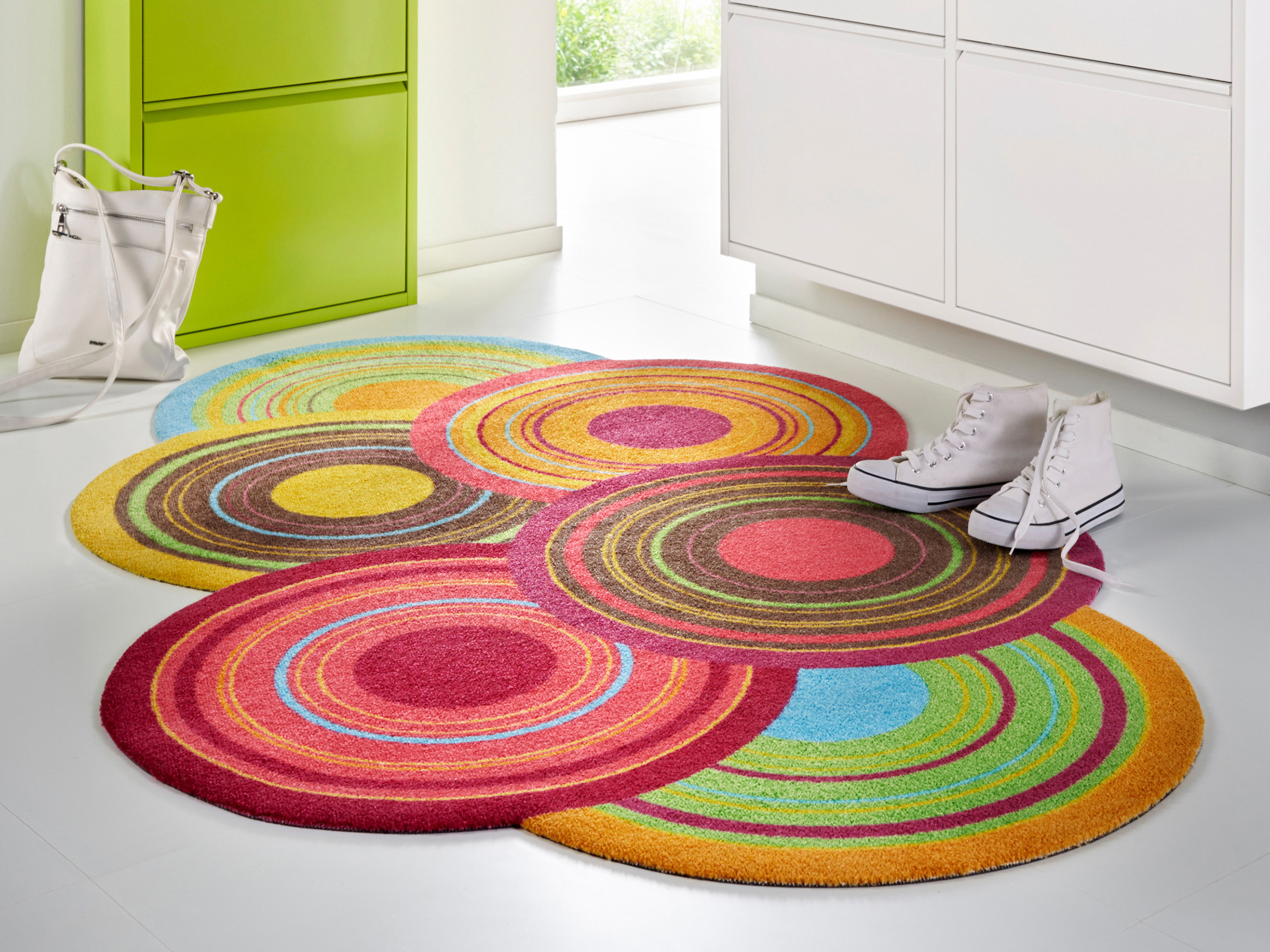 Un tapis coloré avec des motifs circulaires de différentes couleurs est posé sur un sol clair. Une paire de chaussures blanches et un sac blanc sont placés à côté du tapis, tandis qu'une armoire verte est visible en arrière-plan.