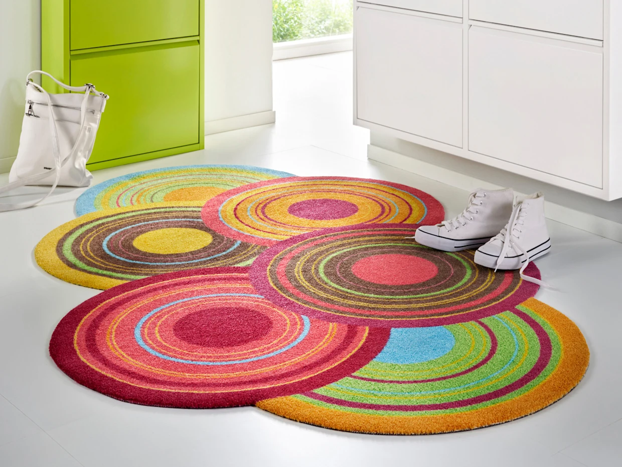 Un tapis coloré avec des motifs circulaires de différentes couleurs est posé sur un sol clair. Une paire de chaussures blanches et un sac blanc sont placés à côté du tapis, tandis qu'une armoire verte est visible en arrière-plan.