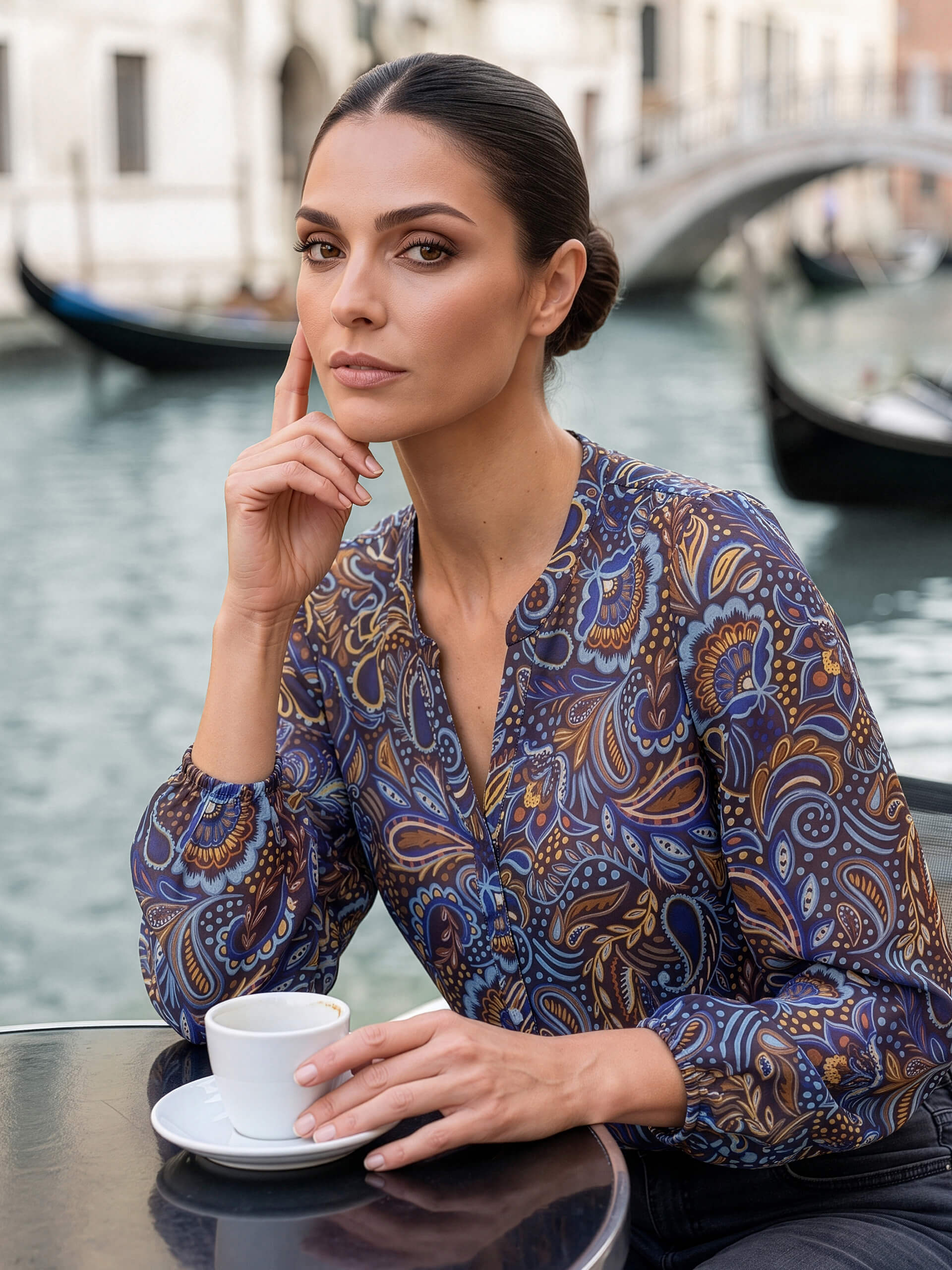 Ein Model sitzt an einem Kanal mit Gondeln und Brücke, trägt eine blau gemusterte Bluse und hält eine Tasse.