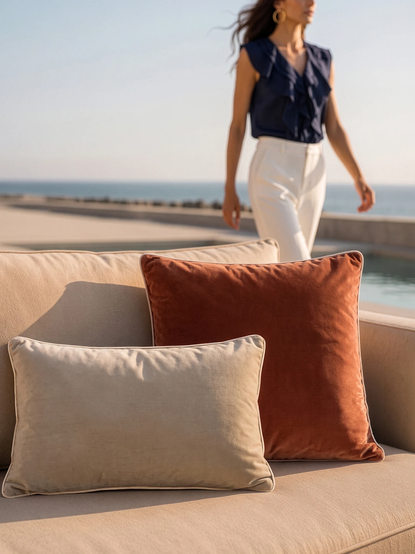 Deux coussins, un beige et un rouille, sont posés sur un canapé clair. En arrière-plan, un modèle en chemisier bleu foncé et pantalon clair marche près de l'eau.
