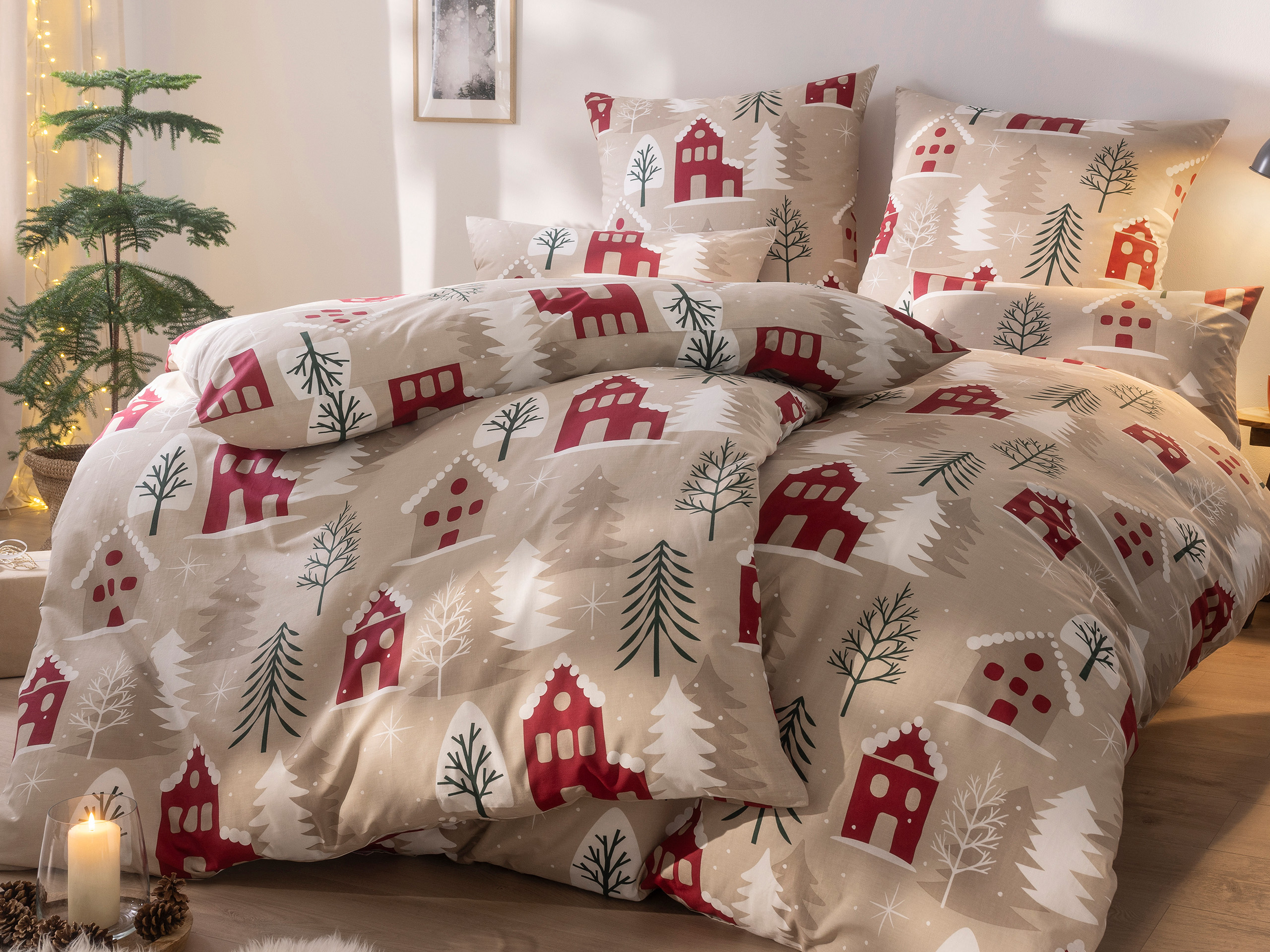 Bettwäsche mit winterlichem Muster aus roten Häusern, Tannenbäumen und Schneeflocken, dekoriert mit Kerzen und Tannenzapfen im Zimmer.