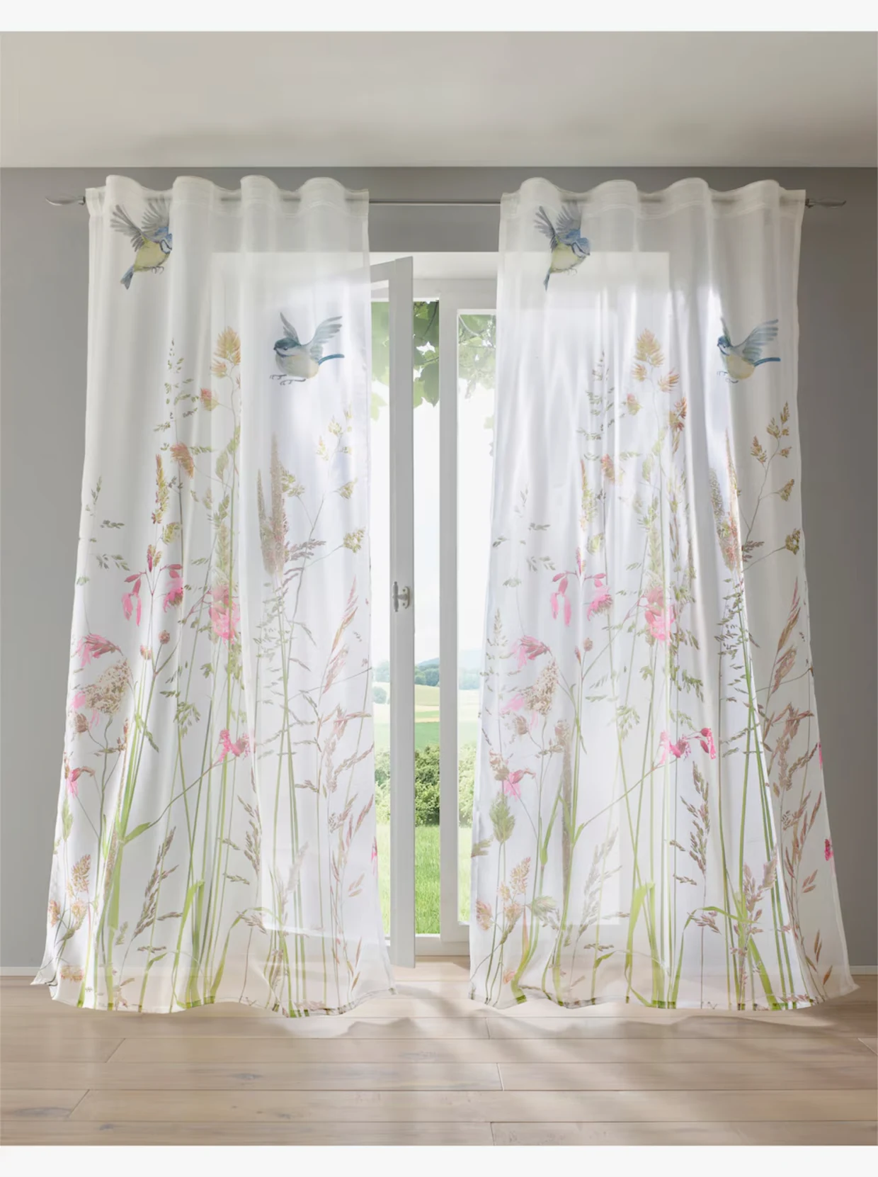 Vor einem Fenster hängen zwei halbdurchsichtige Vorhänge, die mit farbenfrohen Blumenmotiven und Vögeln bedruckt sind. Vor einem Fenster hängen zwei halbdurchsichtige Vorhänge, die mit farbenfrohen Blumenmotiven und Vögeln bedruckt sind.