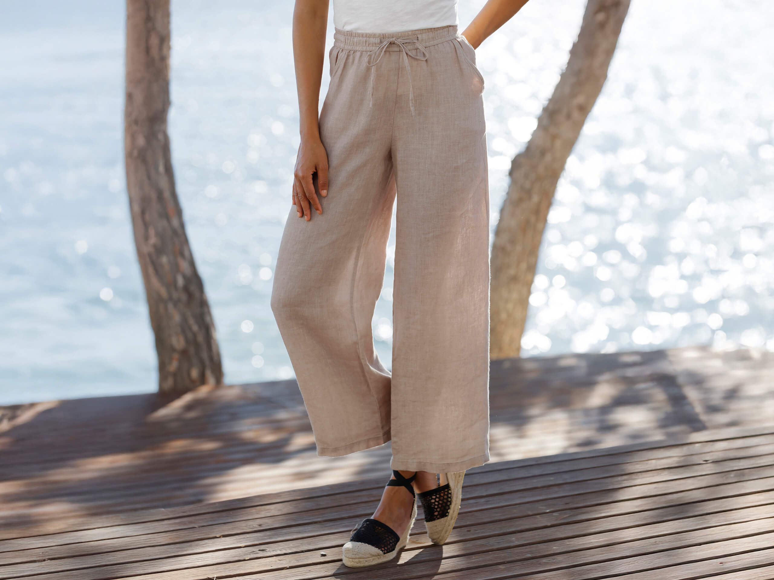Ein Model steht auf einer Holzterrasse am Wasser und trägt eine beige, weite Leinenhose mit Bindeband und schwarze Espadrilles