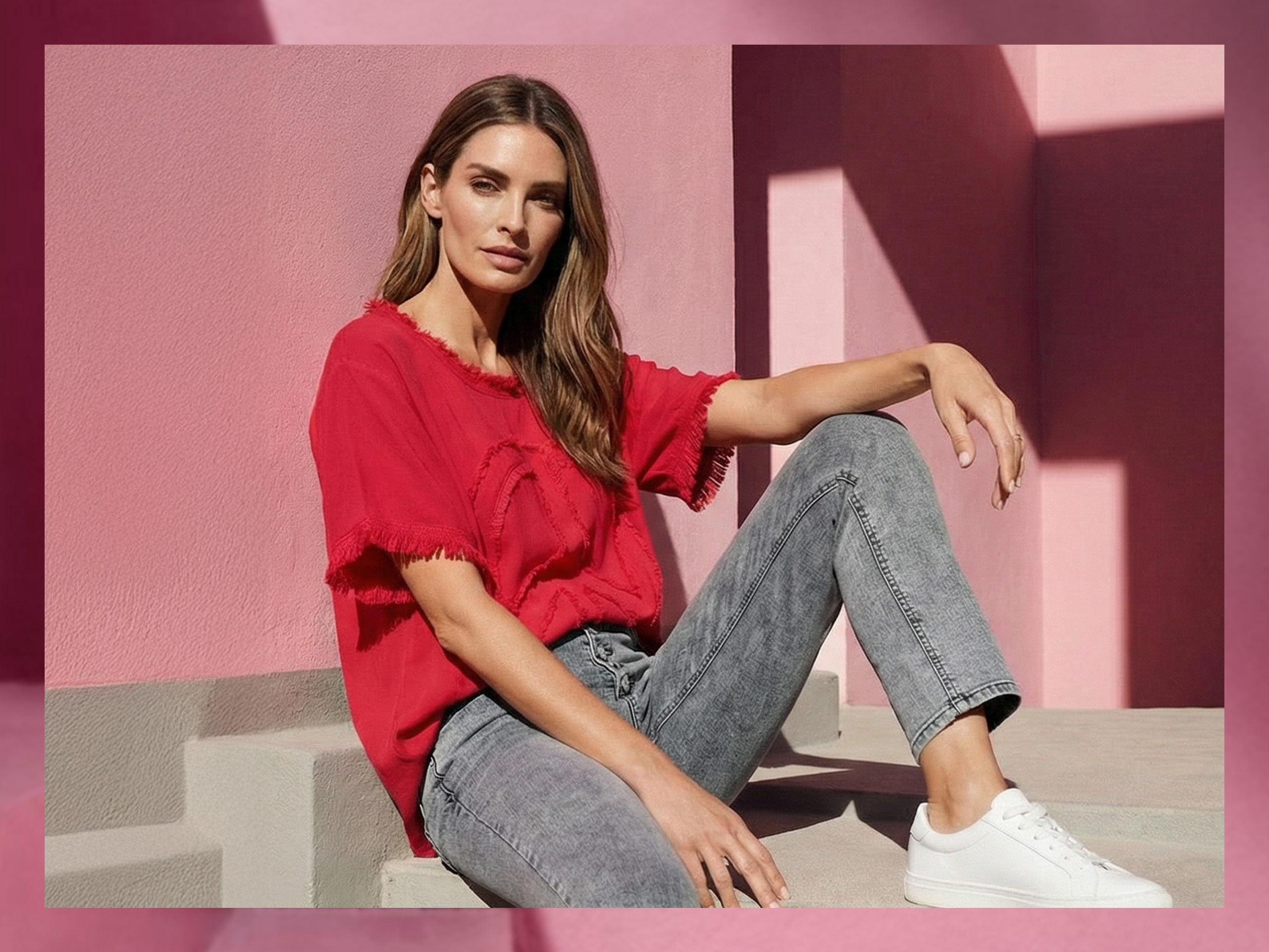 Un modèle est assis sur une marche devant un mur rose, portant un t-shirt rouge, un jean gris et des baskets blanches.