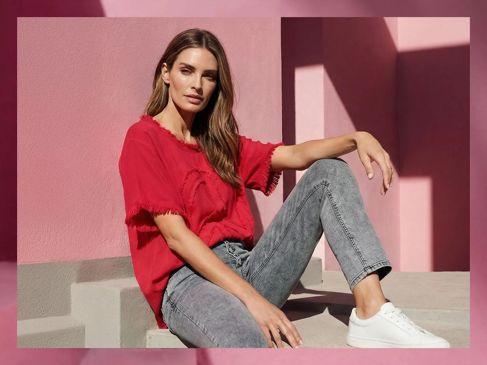 Un modèle est assis sur une marche devant un mur rose, portant un t-shirt rouge, un jean gris et des baskets blanches.