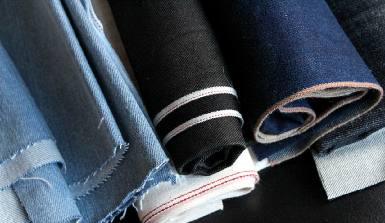 Rollen aus verschiedenfarbigem Denim-Stoff, darunter hellblauer, schwarzer und dunkelblauer Jeansstoff mit sichtbaren Webkanten. Rollen aus verschiedenfarbigem Denim-Stoff, darunter hellblauer, schwarzer und dunkelblauer Jeansstoff mit sichtbaren Webkanten.