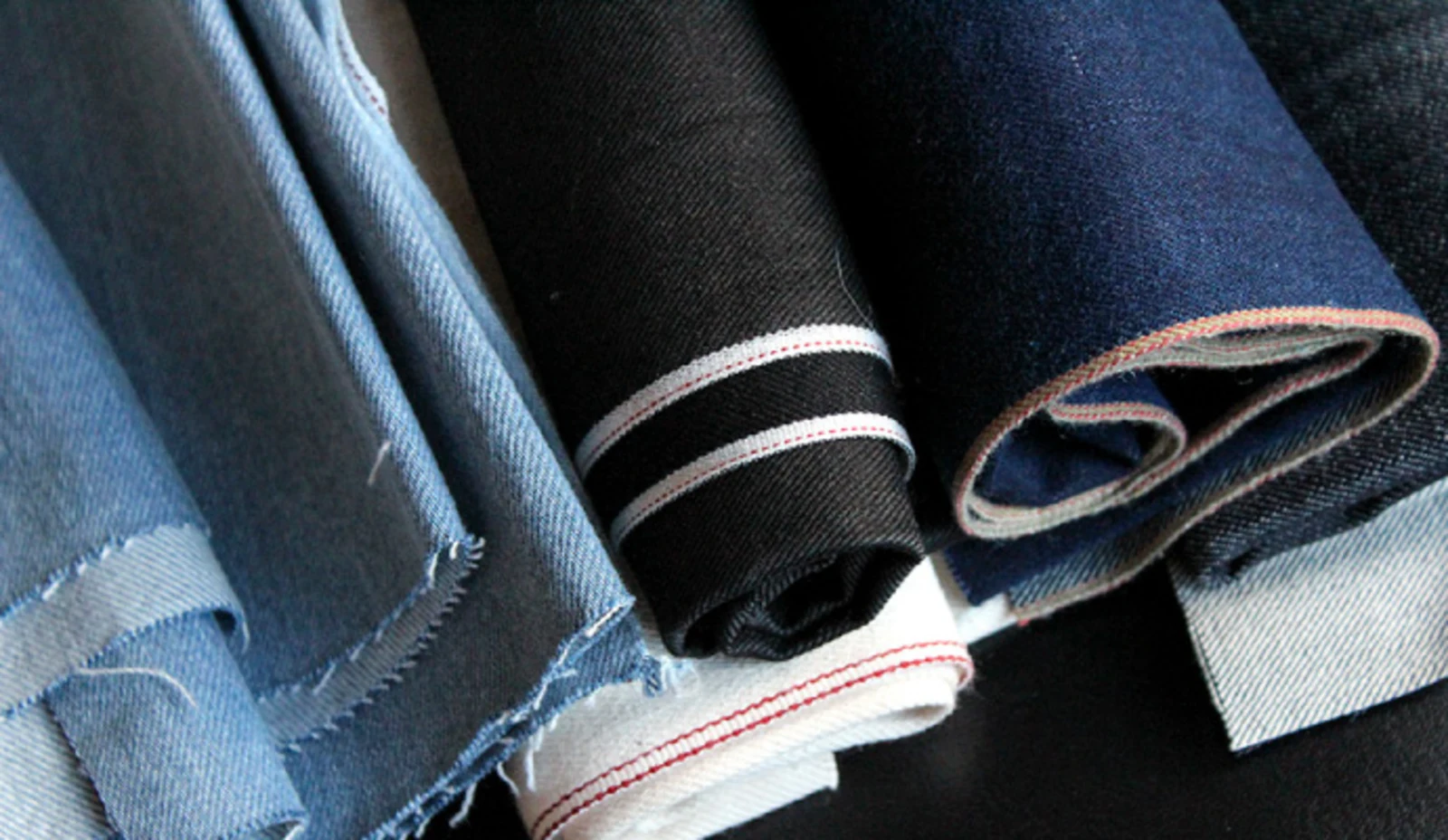 Rollen aus verschiedenfarbigem Denim-Stoff, darunter hellblauer, schwarzer und dunkelblauer Jeansstoff mit sichtbaren Webkanten. Rollen aus verschiedenfarbigem Denim-Stoff, darunter hellblauer, schwarzer und dunkelblauer Jeansstoff mit sichtbaren Webkanten.