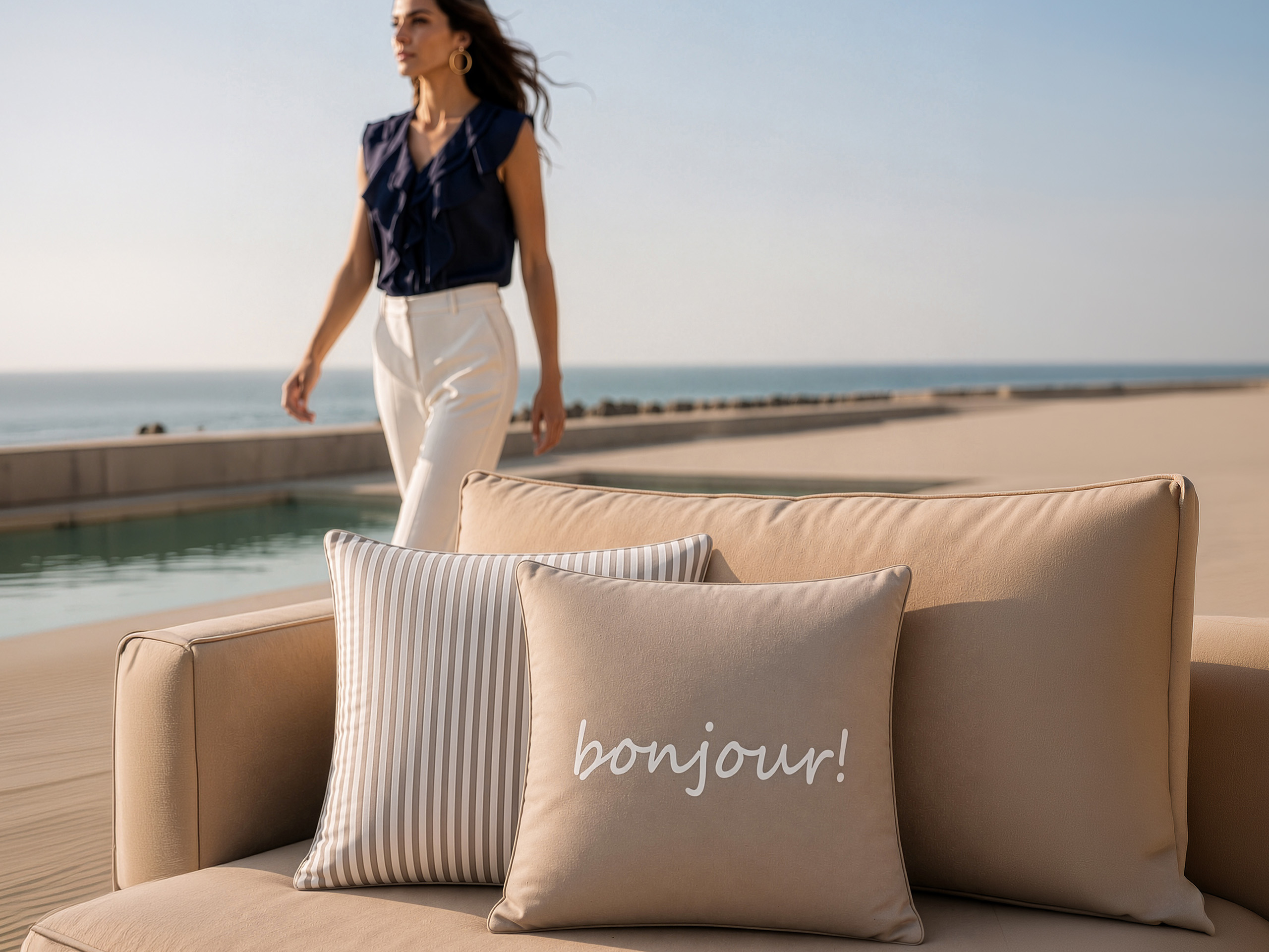 Twee kussens liggen op een beige bank, één gestreept, de ander met de tekst 'bonjour!'. Op de achtergrond loopt een model met een donkerblauwe blouse en lichte broek langs het water.