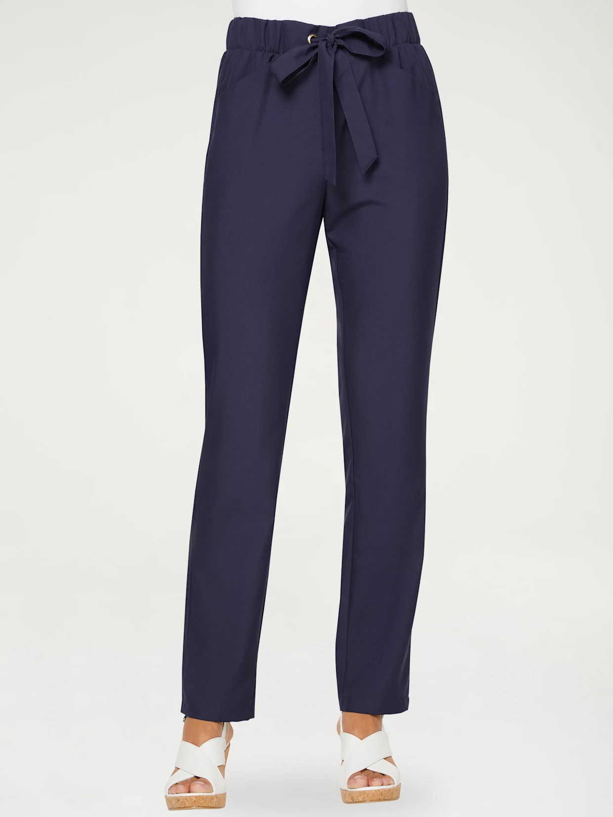 Pantalon bleu marine pour femme avec taille élastique et ceinture nouée, porté avec des sandales blanches compensées. Pantalon bleu marine pour femme avec taille élastique et ceinture nouée, porté avec des sandales blanches compensées.