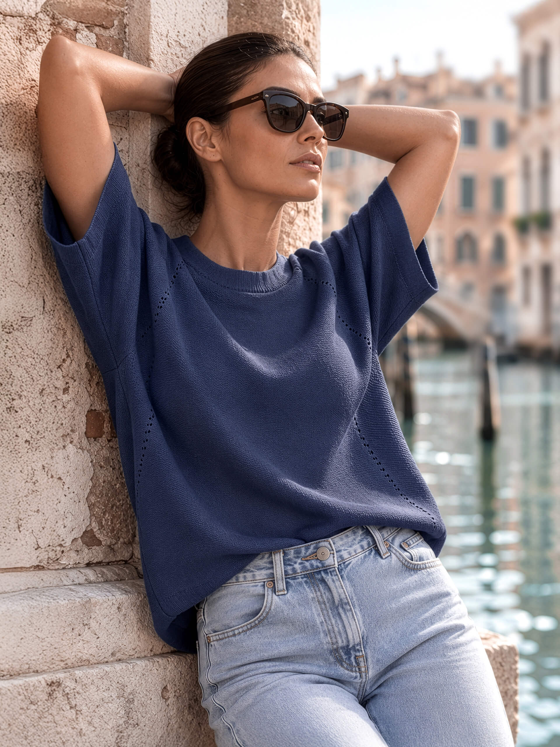 Ein Model in blauem Oberteil und Jeans lehnt an einer Steinwand an einem Kanal und trägt eine Sonnenbrille.