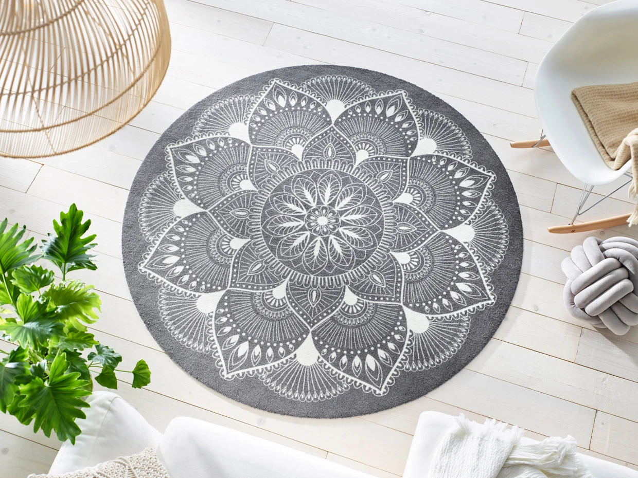 Ein heller Raum ist mit einem runden Teppich in Grau und Weiß dekoriert, der ein detailliertes Mandala-Muster zeigt. Umgeben von einem bequemen Stuhl und grünen Pflanzen schafft der Teppich eine entspannende Atmosphäre auf dem hellen Holzboden.