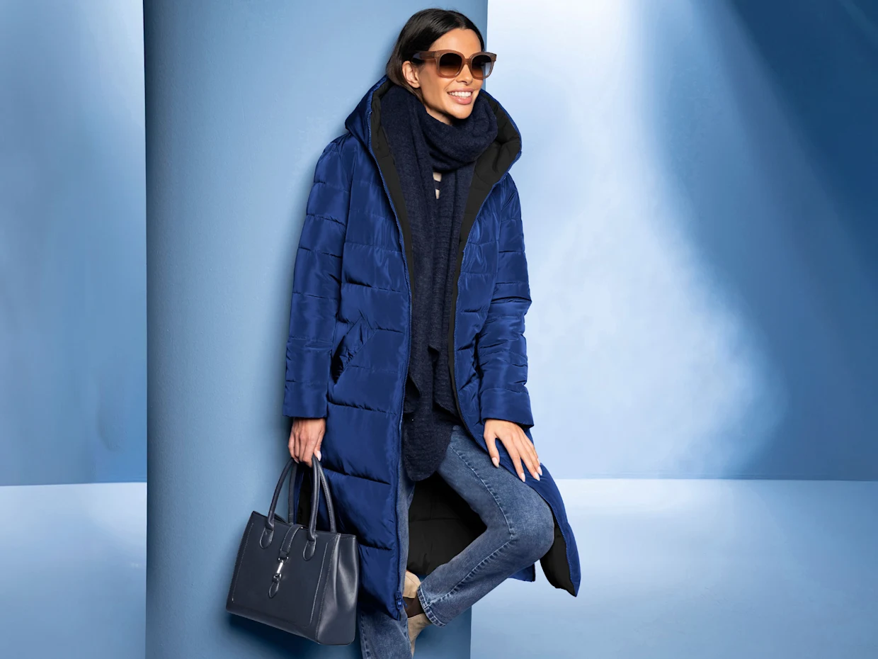 Un mannequin porte un long manteau matelassé bleu foncé, une écharpe, un jean et tient un sac à main bleu. Fond dans les tons bleus. Un mannequin porte un long manteau matelassé bleu foncé, une écharpe, un jean et tient un sac à main bleu. Fond dans les tons bleus.