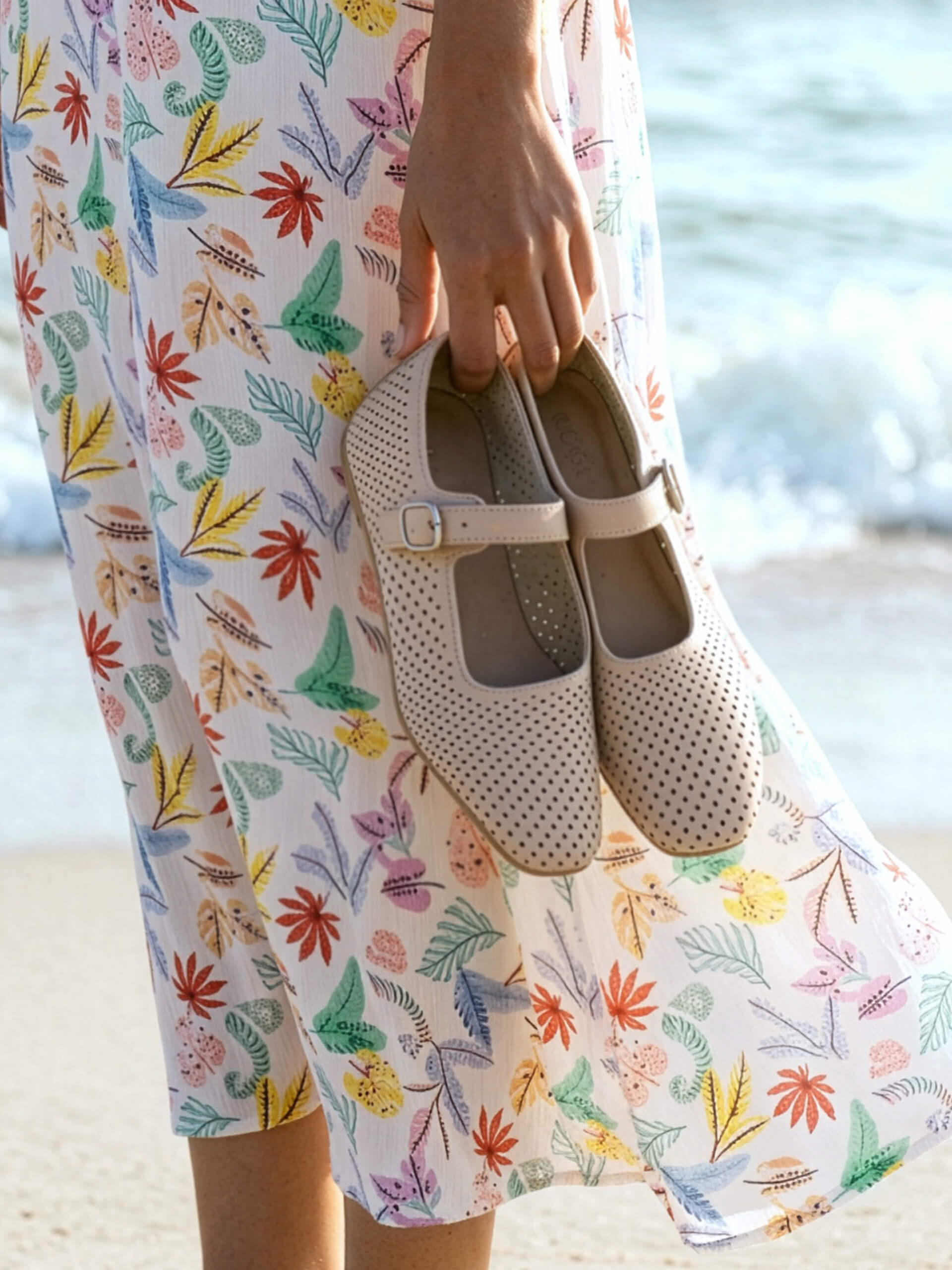 Ein Model steht am Strand in einem geblümten Rock und hält beigefarbene, gelochte Ballerinas in der Hand.