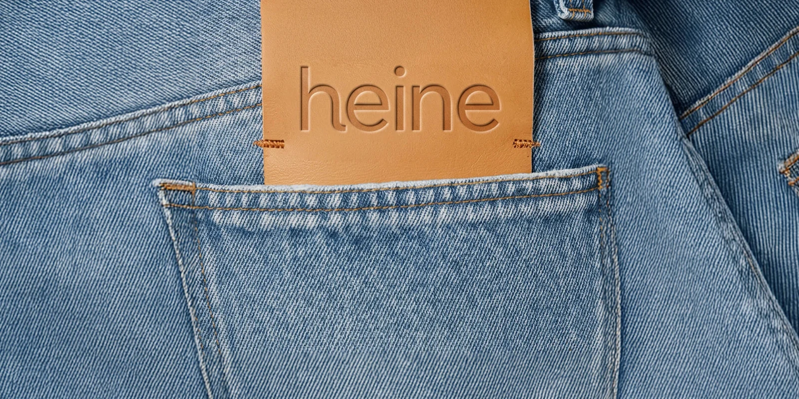 Detailaufnahme einer hellblauen Jeans mit braunem Lederetikett in der Tasche. Auf dem Etikett steht: 'heine'.