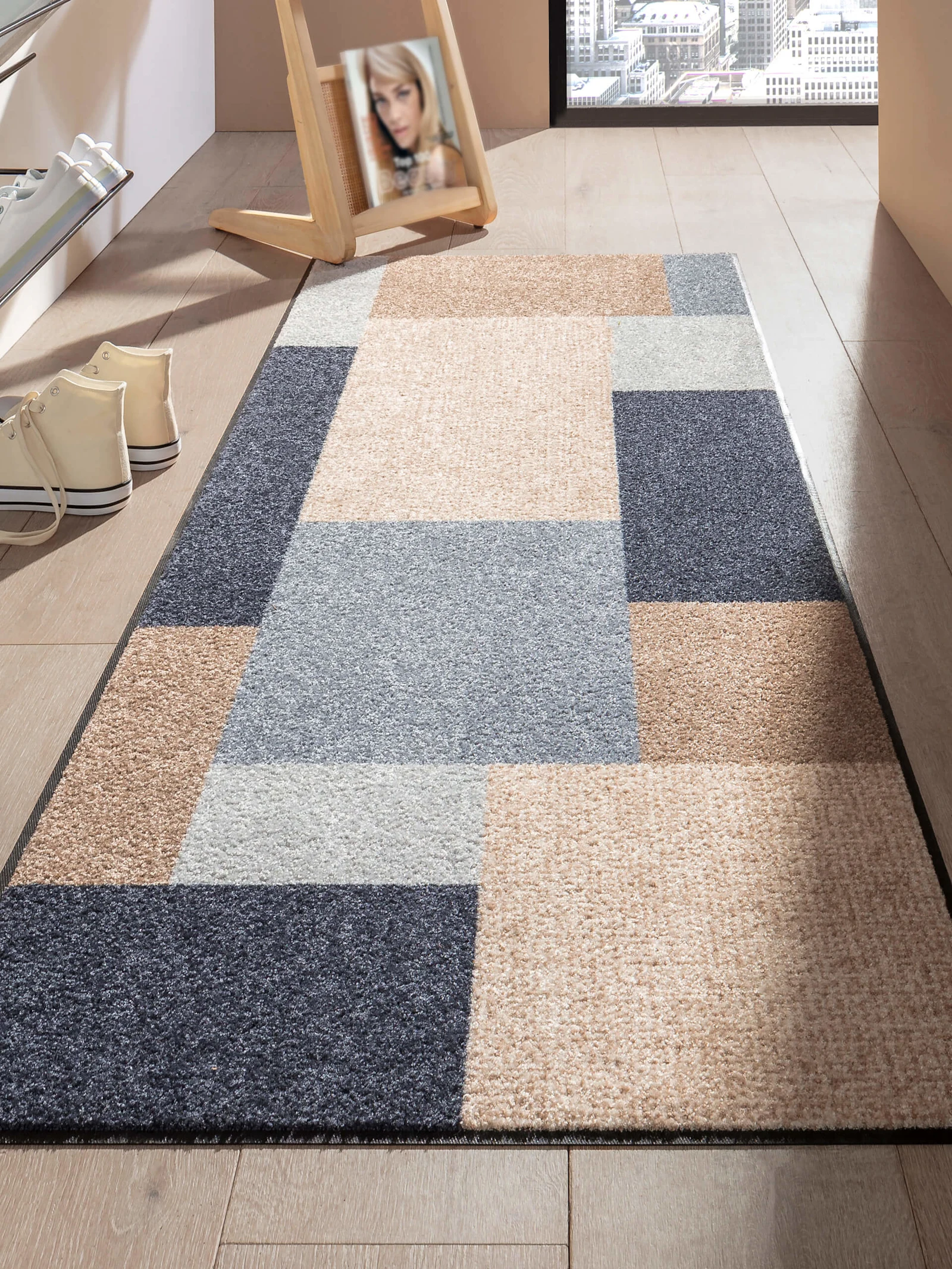 Un tapis à motif géométrique en tons beige, gris et bleu est posé sur un sol en bois clair, à côté deux paires de chaussures et un miroir.