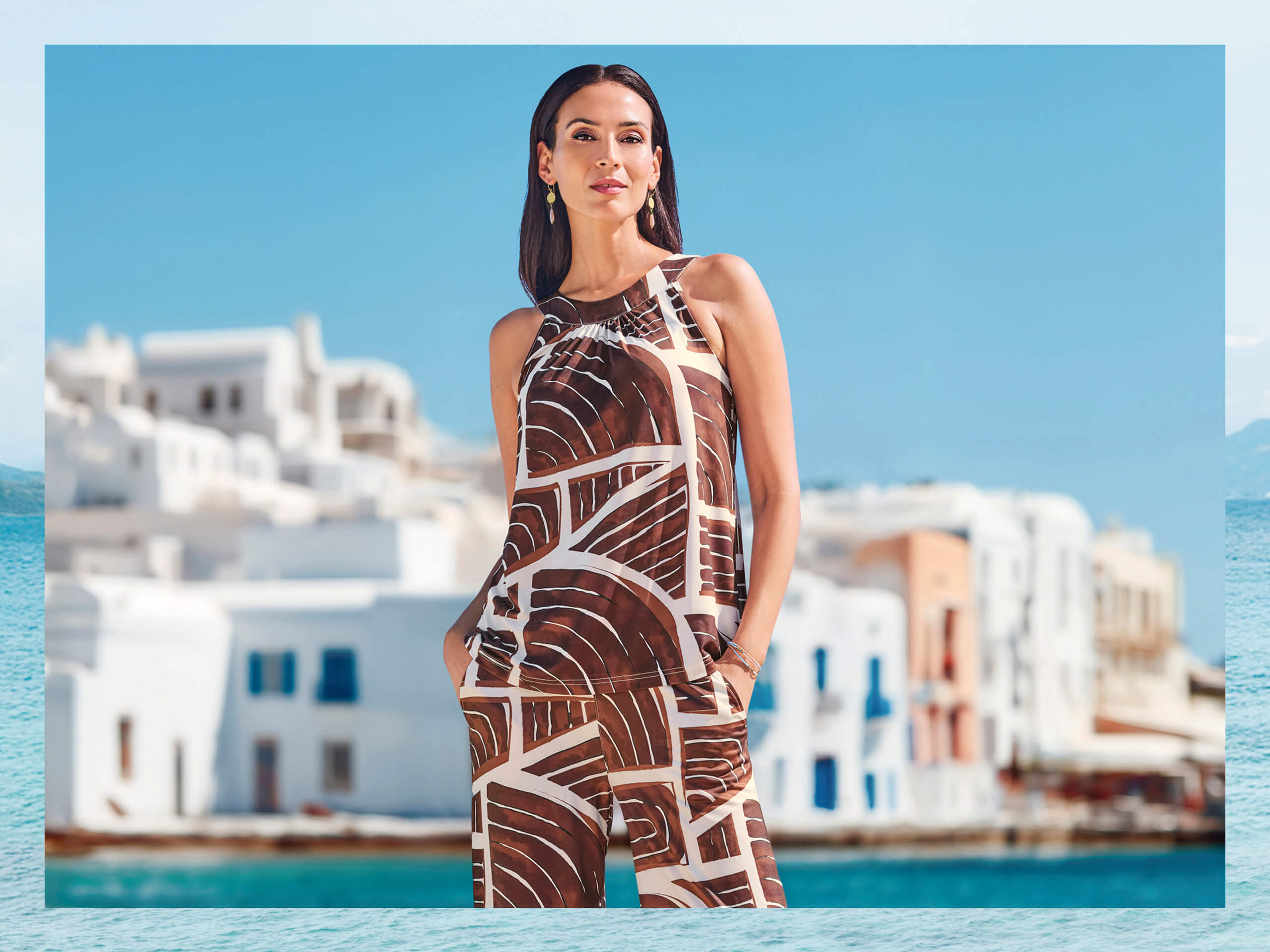 Ein Model steht am Wasser vor weißen Häusern, trägt ein ärmelloses Top und weite Hose mit braun-weißem abstraktem Muster.