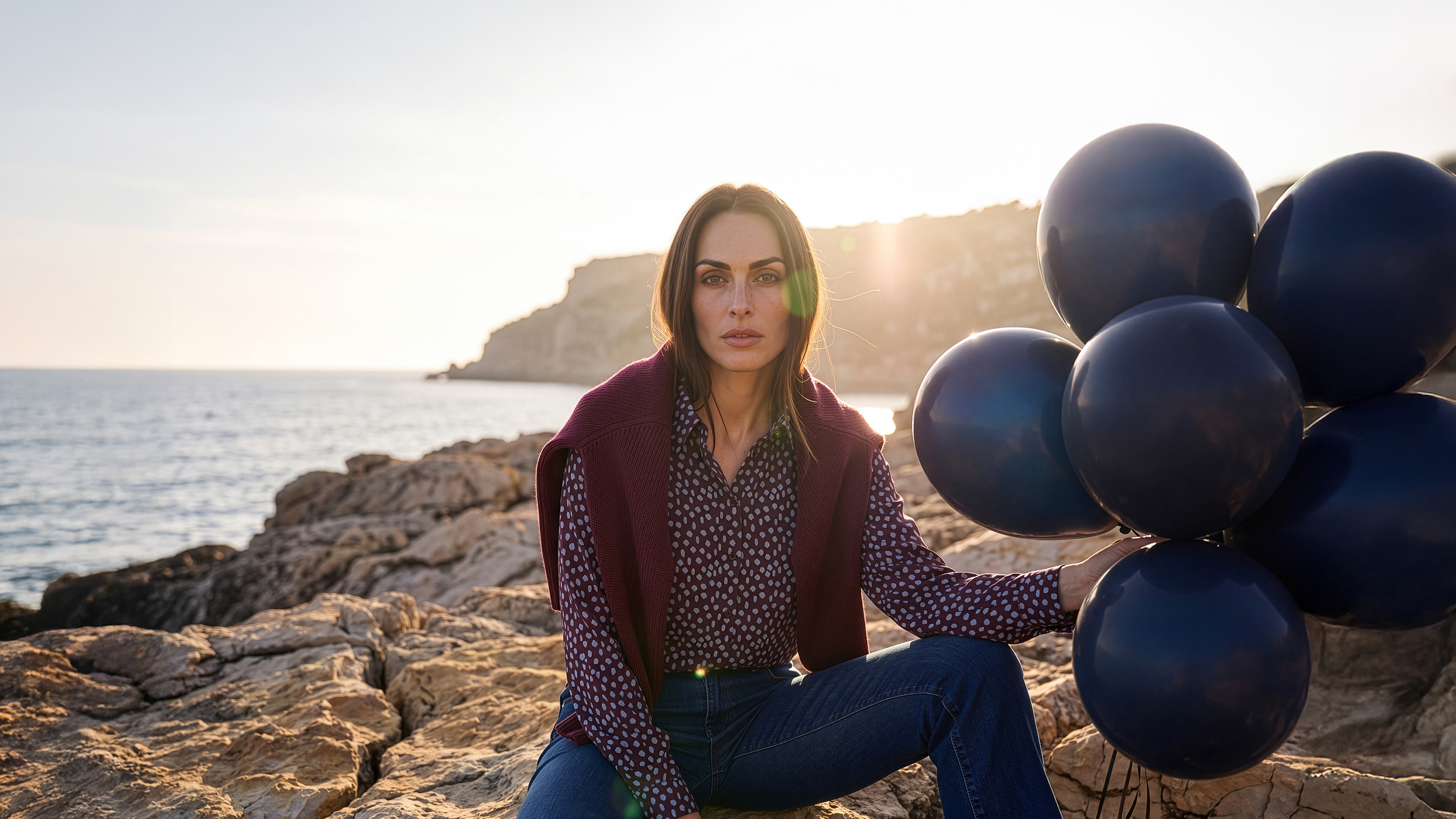 Ein Model sitzt auf einem Felsen am Meer. Es trägt eine gemusterte Bluse, eine Jeans und einen Pullover über den Schultern. 