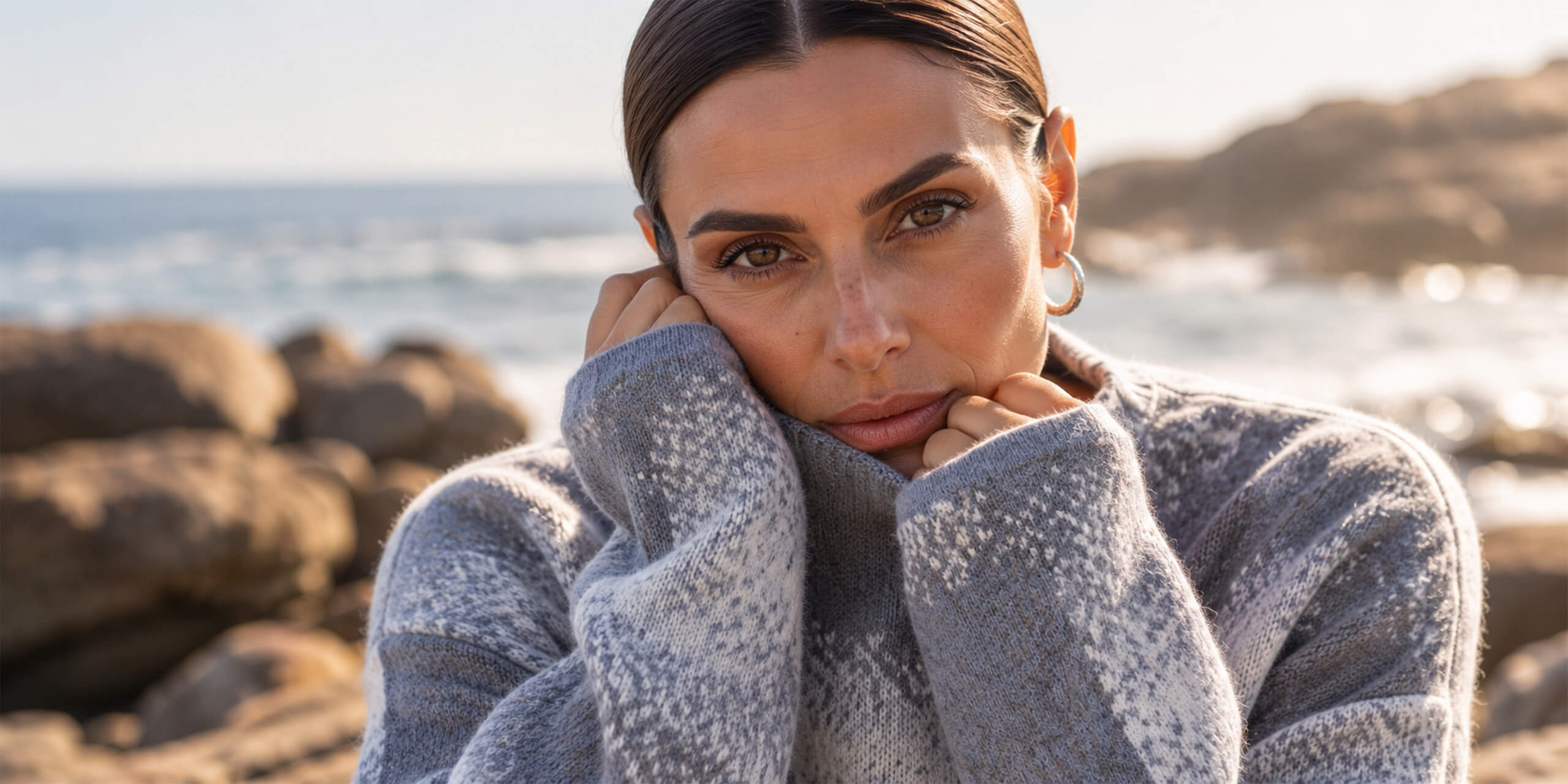 Ein Model in einem grauen Strickpullover mit Muster. Sie hat die Hände am Gesicht und blickt an der Küste in die Kamera.