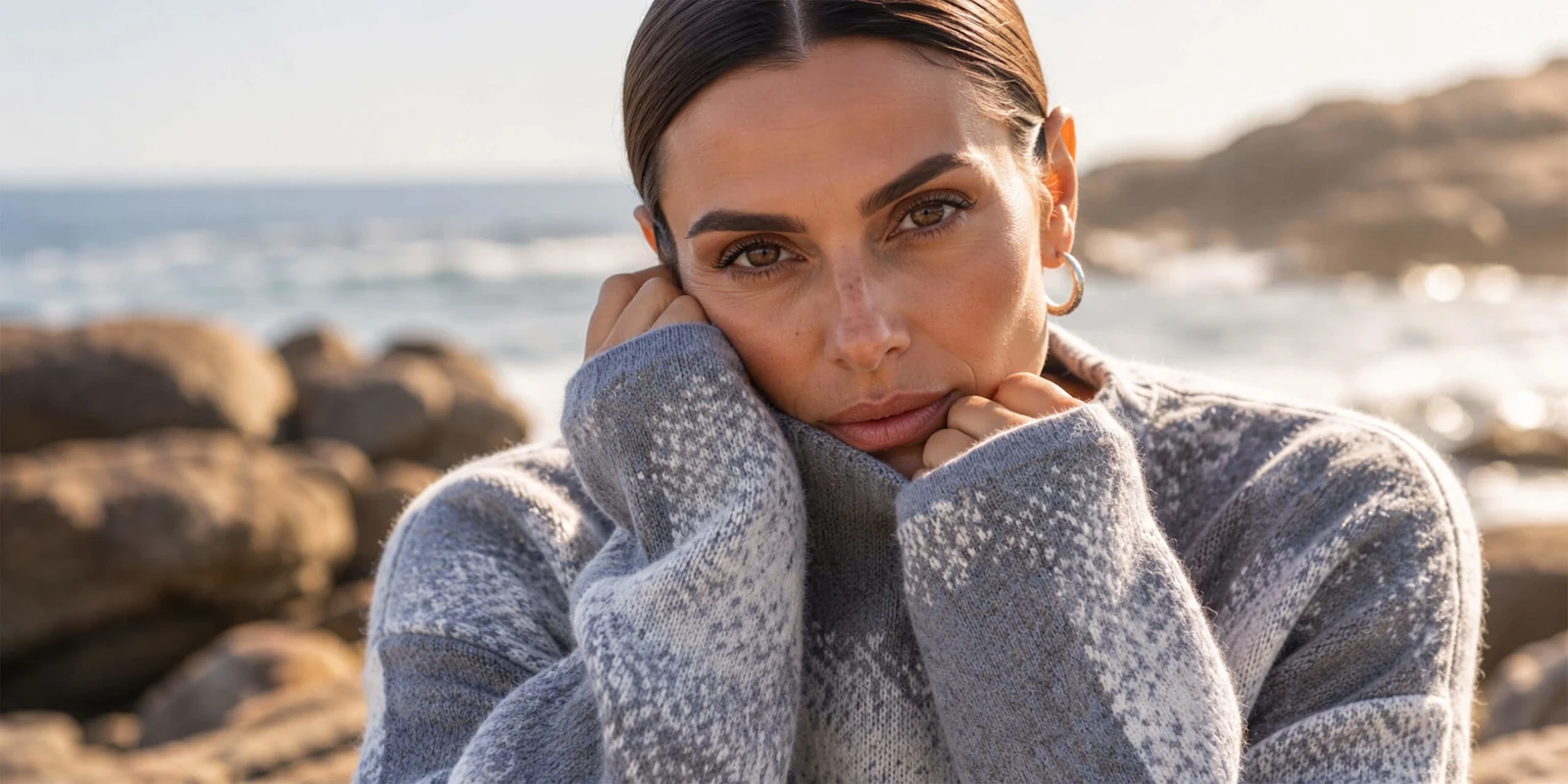 Ein Model in einem grauen Strickpullover mit Muster. Sie hat die Hände am Gesicht und blickt an der Küste in die Kamera.