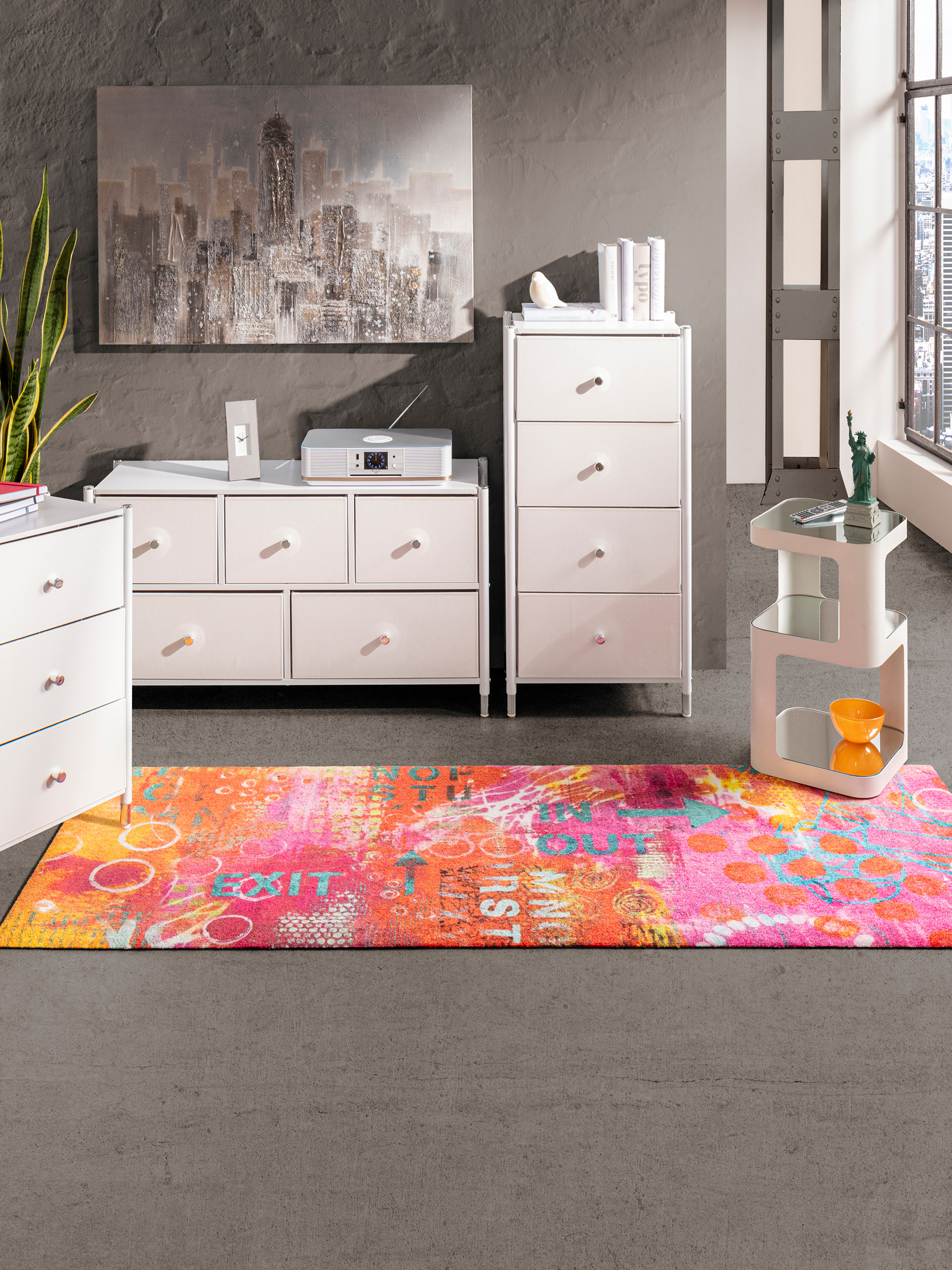 Ein Wohnzimmer ist mit weissen Möbeln und einem bunten Teppich mit Schriftzügen und Kreismustern ausgestattet. An der Wand hängt ein grosses Bild einer Stadtlandschaft, und durch die Fenster fällt viel Licht in den Raum.