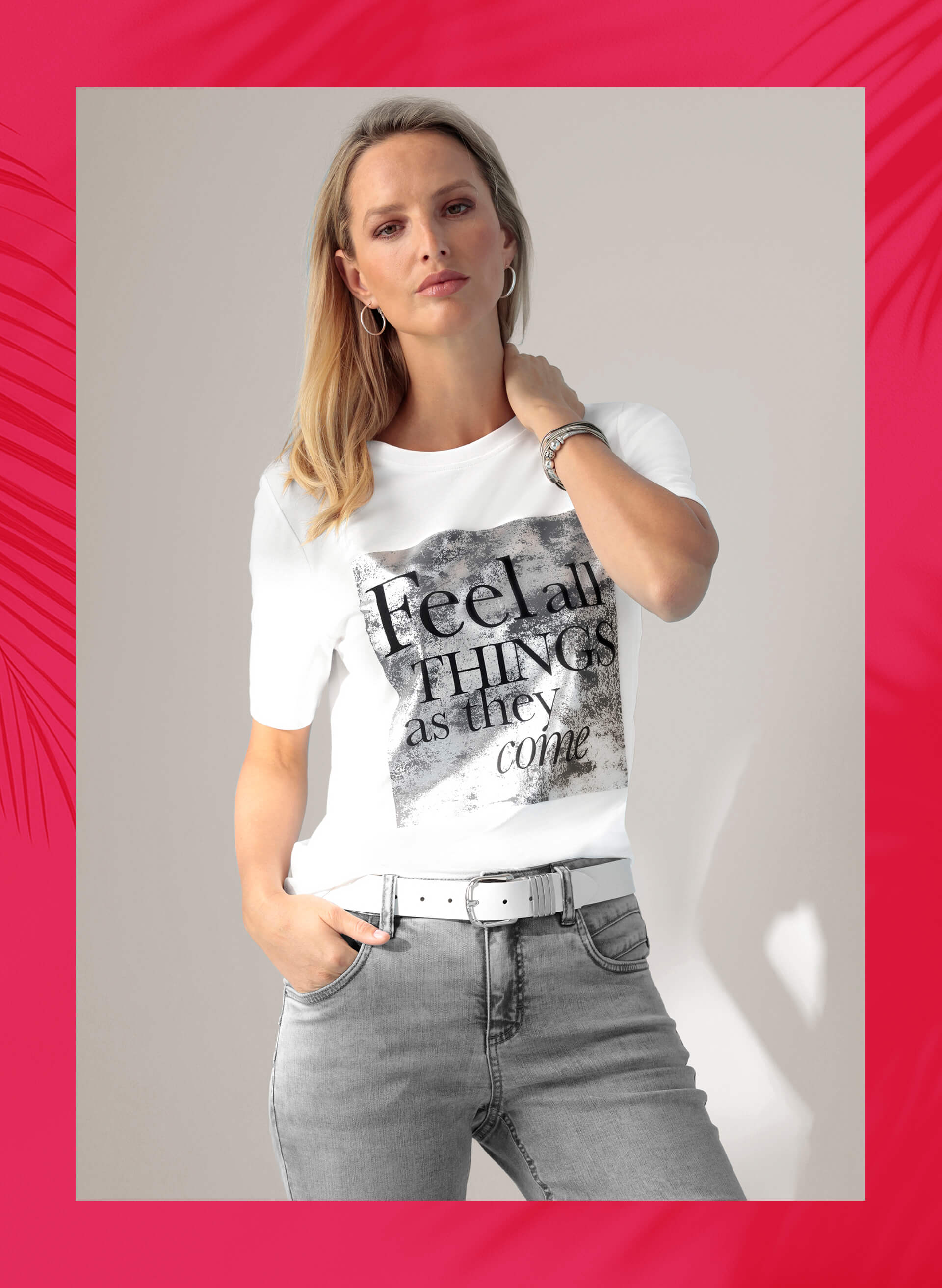Ein Model trägt weißes T-Shirt mit schwarzem Schriftprint und graue Jeans, eine Hand in der Hosentasche.