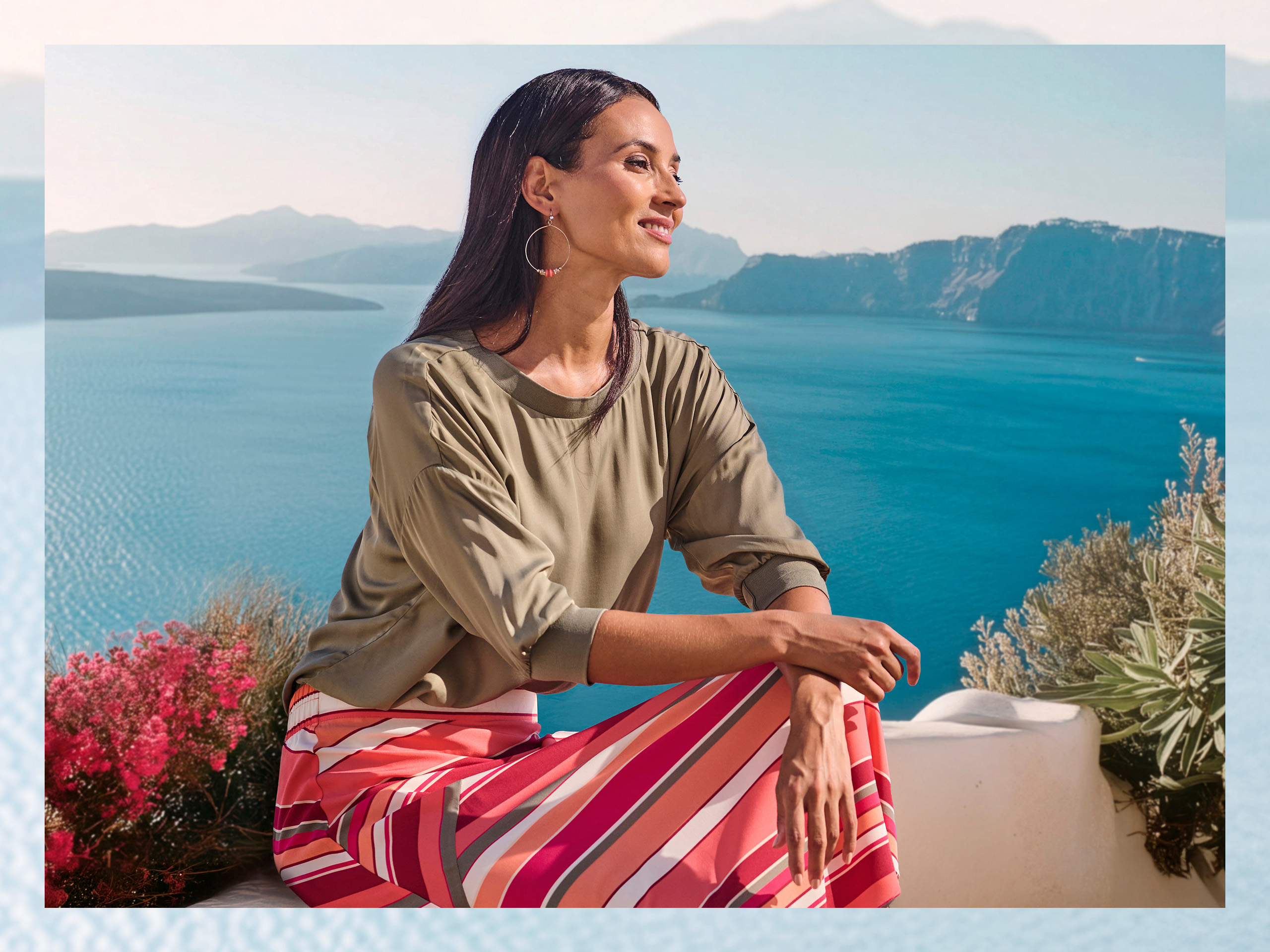 Un modèle porte un pull vert olive à manches 3/4 et une jupe rayée colorée, assis devant un paysage marin avec des montagnes.
