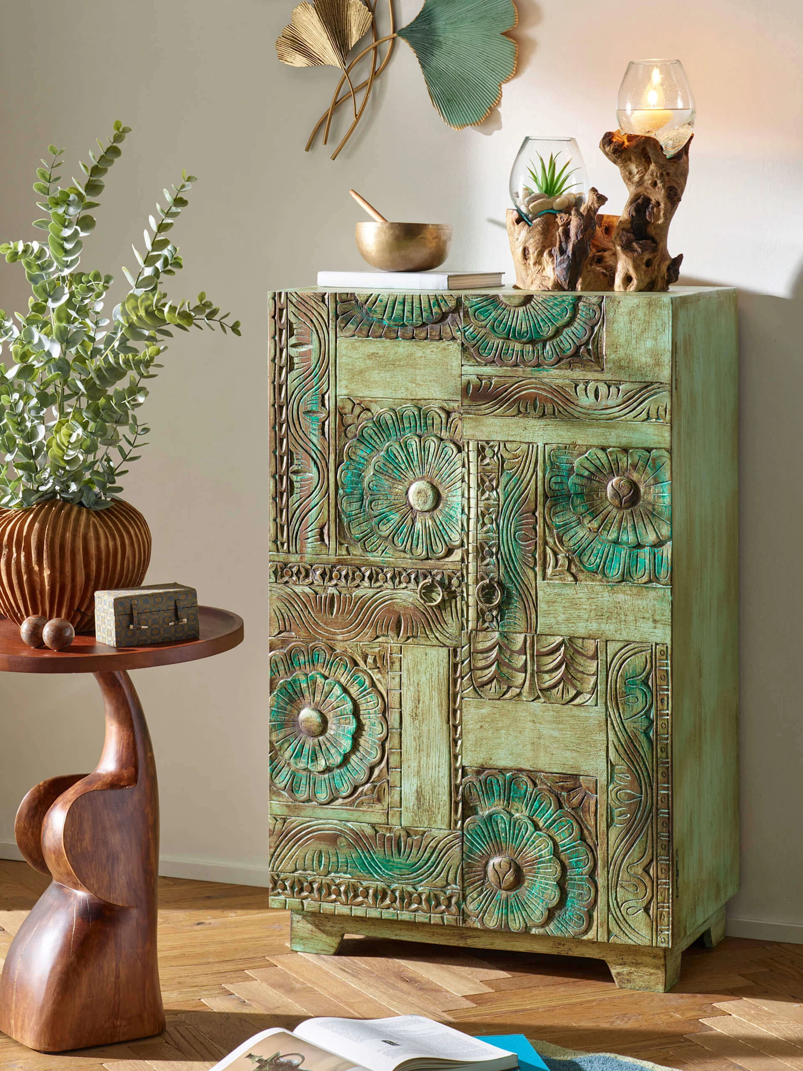 Une armoire verte finement décorée se trouve à côté d'une petite table d'appoint marron avec un vase et des plantes, dans une pièce lumineuse avec fenêtre.