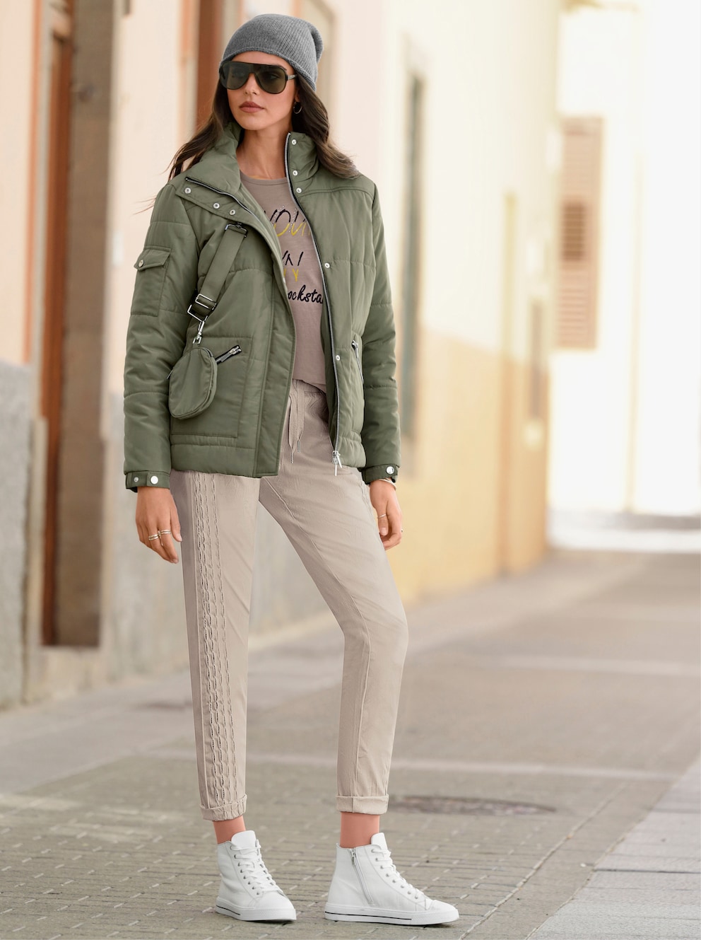 Model steht auf der Staße und trägt eine khakifarbene Steppjacke Jacke. Dazu trägt sie eine beige Hose, einen braunen Pullover und weiße Sneaker. 