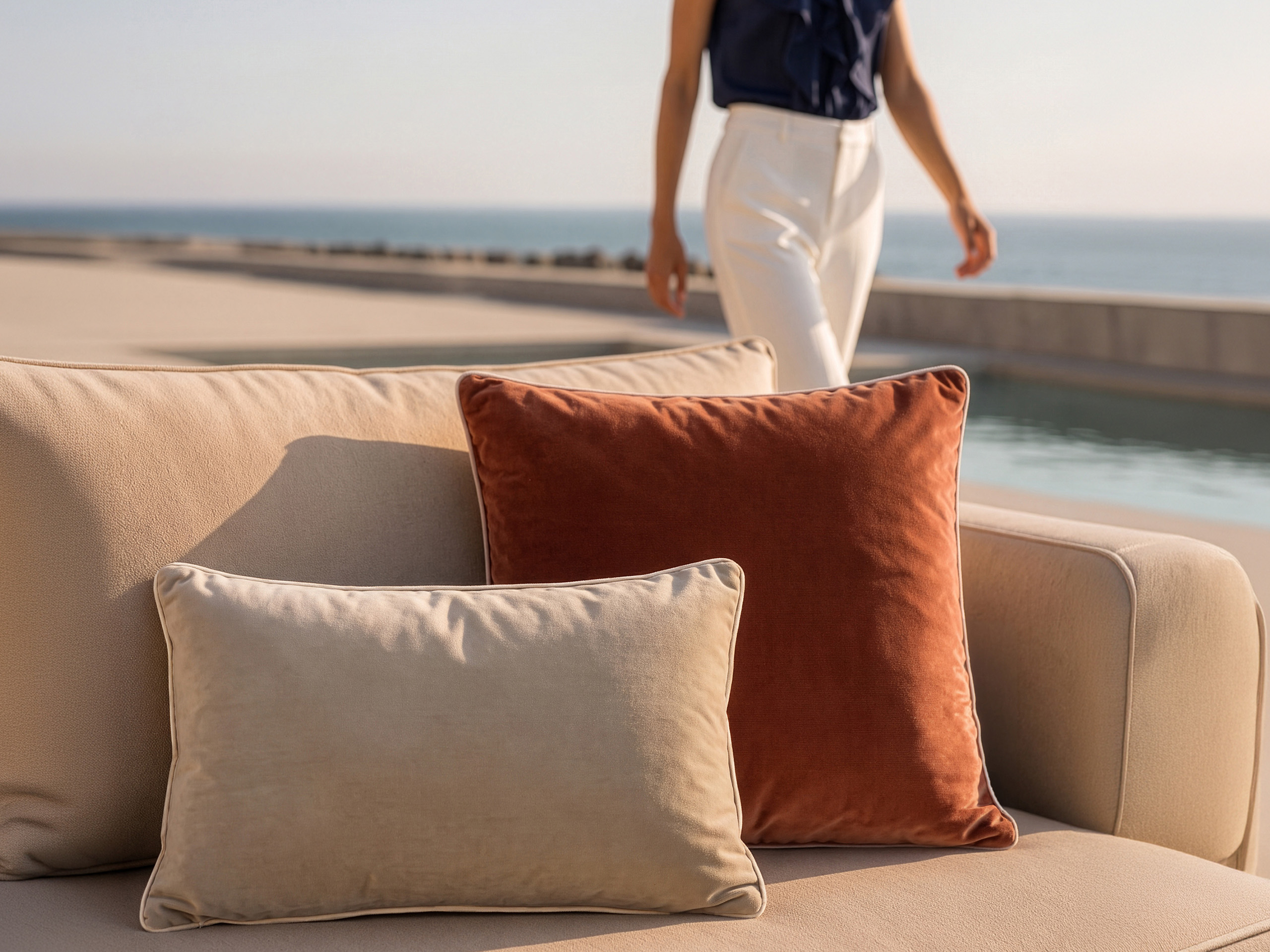 Zwei Kissen, eines beige und eines rostfarben, liegen auf einem hellen Sofa. Im Hintergrund geht ein Model mit dunkelblauer Bluse und heller Hose am Wasser entlang.