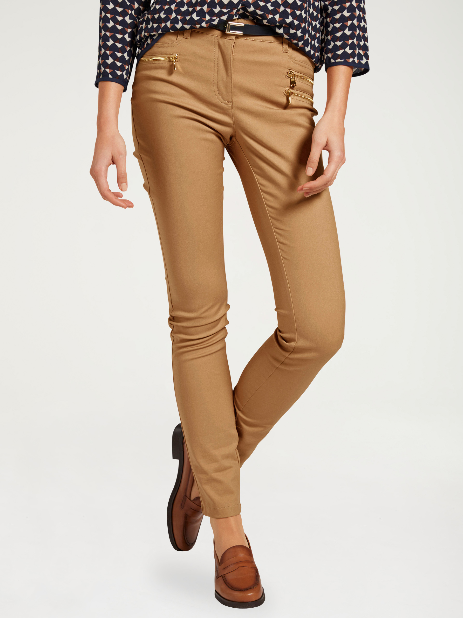 Pantalon pour femme en camel avec une coupe étroite, des fermetures éclair décoratives dorées et des chaussures brunes combinées.
