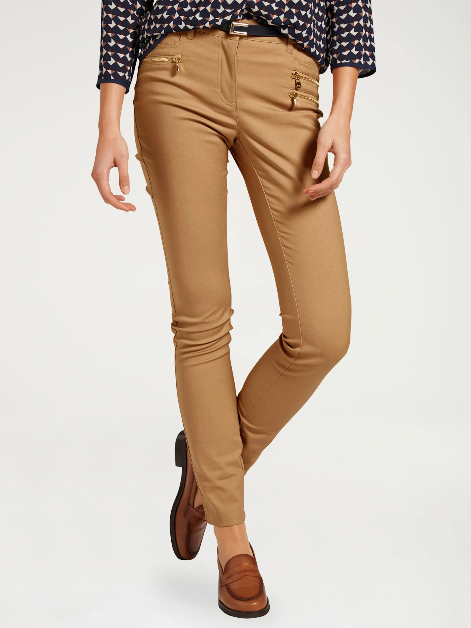 Pantalon pour femme en camel avec une coupe étroite, des fermetures éclair décoratives dorées et des chaussures brunes combinées.