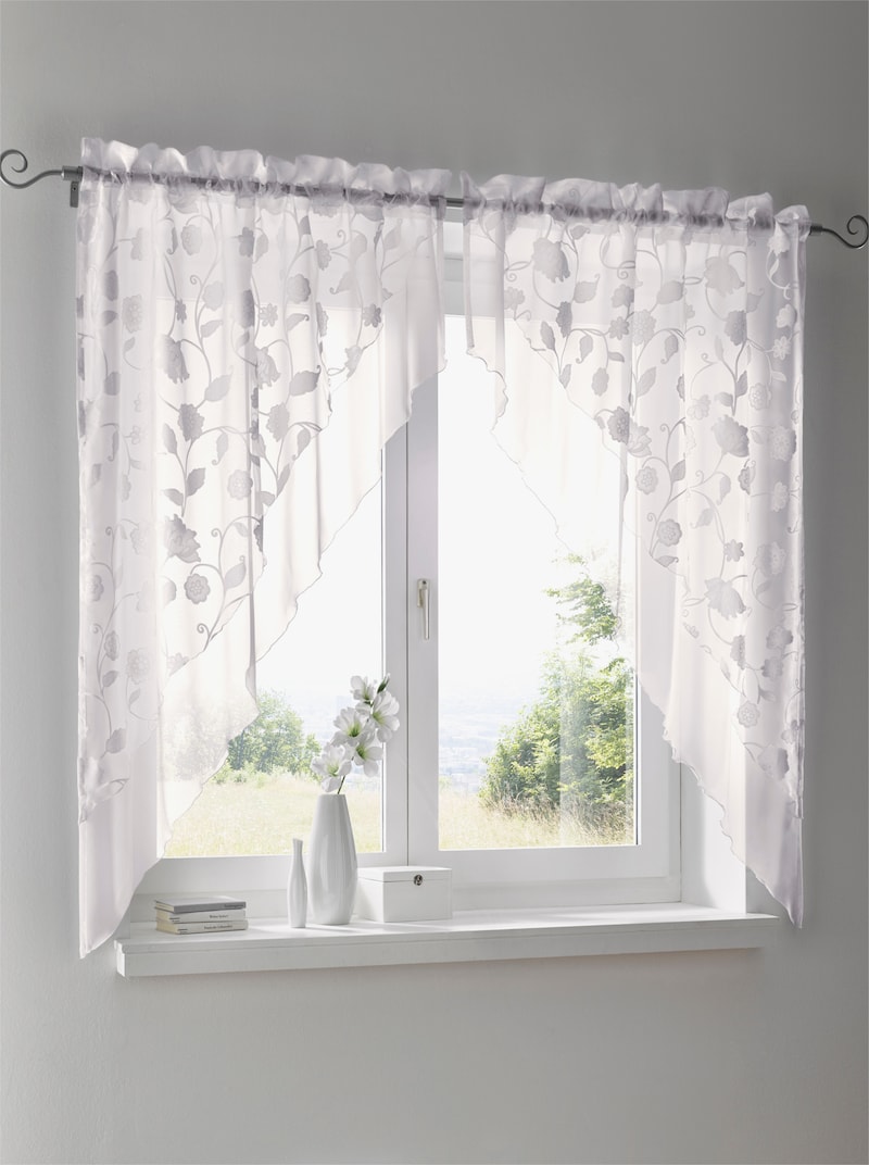 Weiße Gardinen mit floralem Muster an einem Fenster, darunter eine Fensterbank mit einer weißen Vase, Blumen und Dekoration.