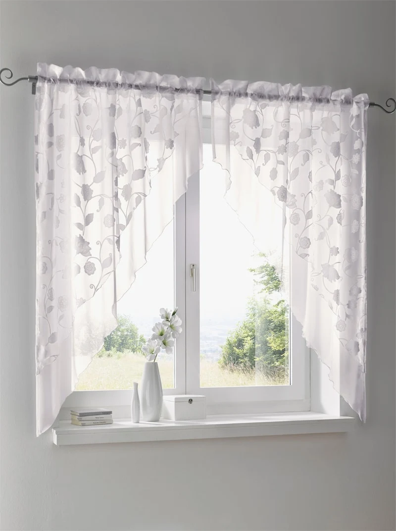 Weiße Gardinen mit floralem Muster an einem Fenster, darunter eine Fensterbank mit einer weißen Vase, Blumen und Dekoration. Weiße Gardinen mit floralem Muster an einem Fenster, darunter eine Fensterbank mit einer weißen Vase, Blumen und Dekoration.