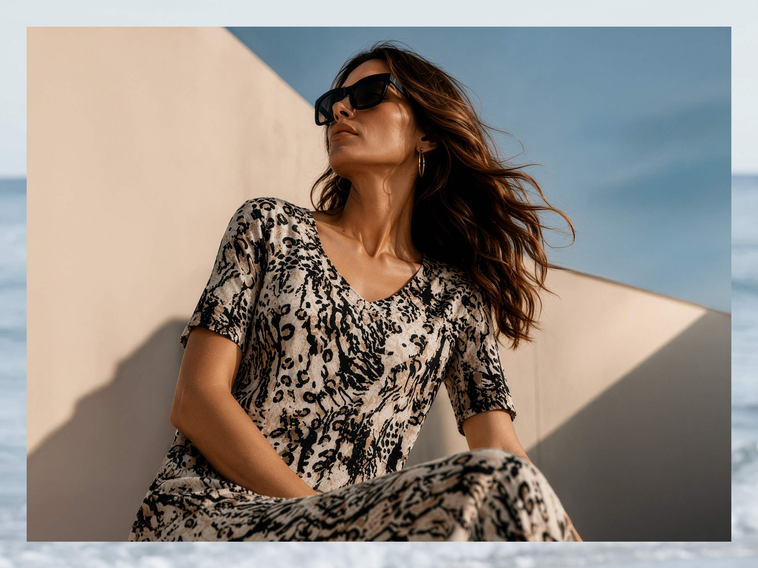 Ein Model trägt ein fließendes, kurzärmeliges Kleid mit Leopardenmuster und Sonnenbrille, posiert vor einer hellen Wand bei blauem Himmel.