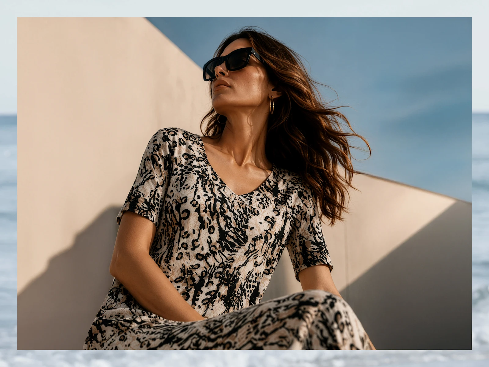 Ein Model trägt ein fließendes, kurzärmeliges Kleid mit Leopardenmuster und Sonnenbrille, posiert vor einer hellen Wand bei blauem Himmel.