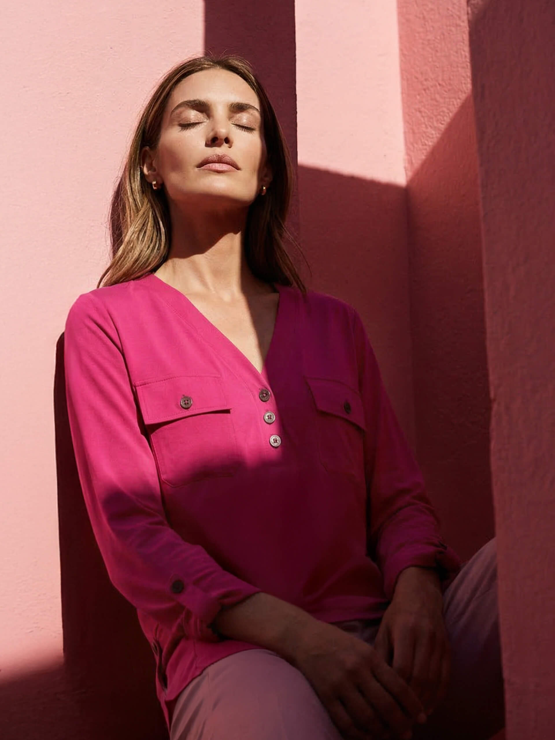 Een model zit voor roze muren en draagt een roze blouse met V-hals en knopen, met opgerolde mouwen.