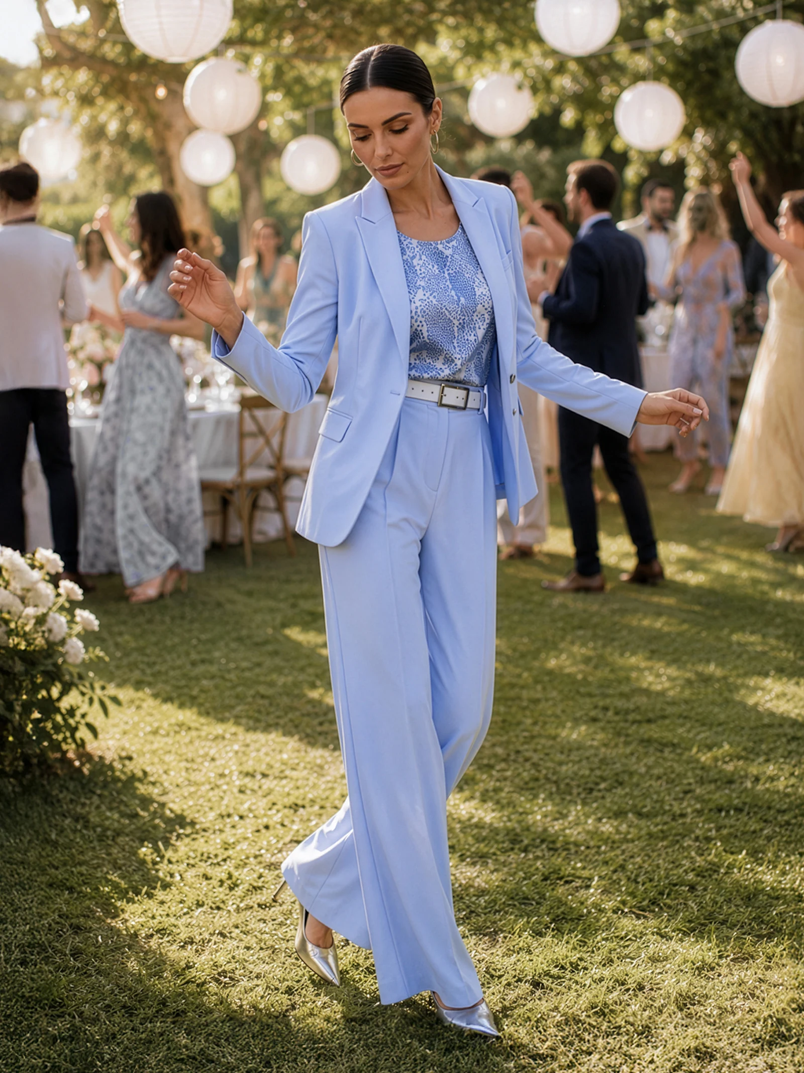 Un mannequin porte un tailleur-pantalon bleu clair avec une ceinture assortie et un top à motifs, se tenant lors d'une garden-party avec des invités et des lampions blancs en arrière-plan.