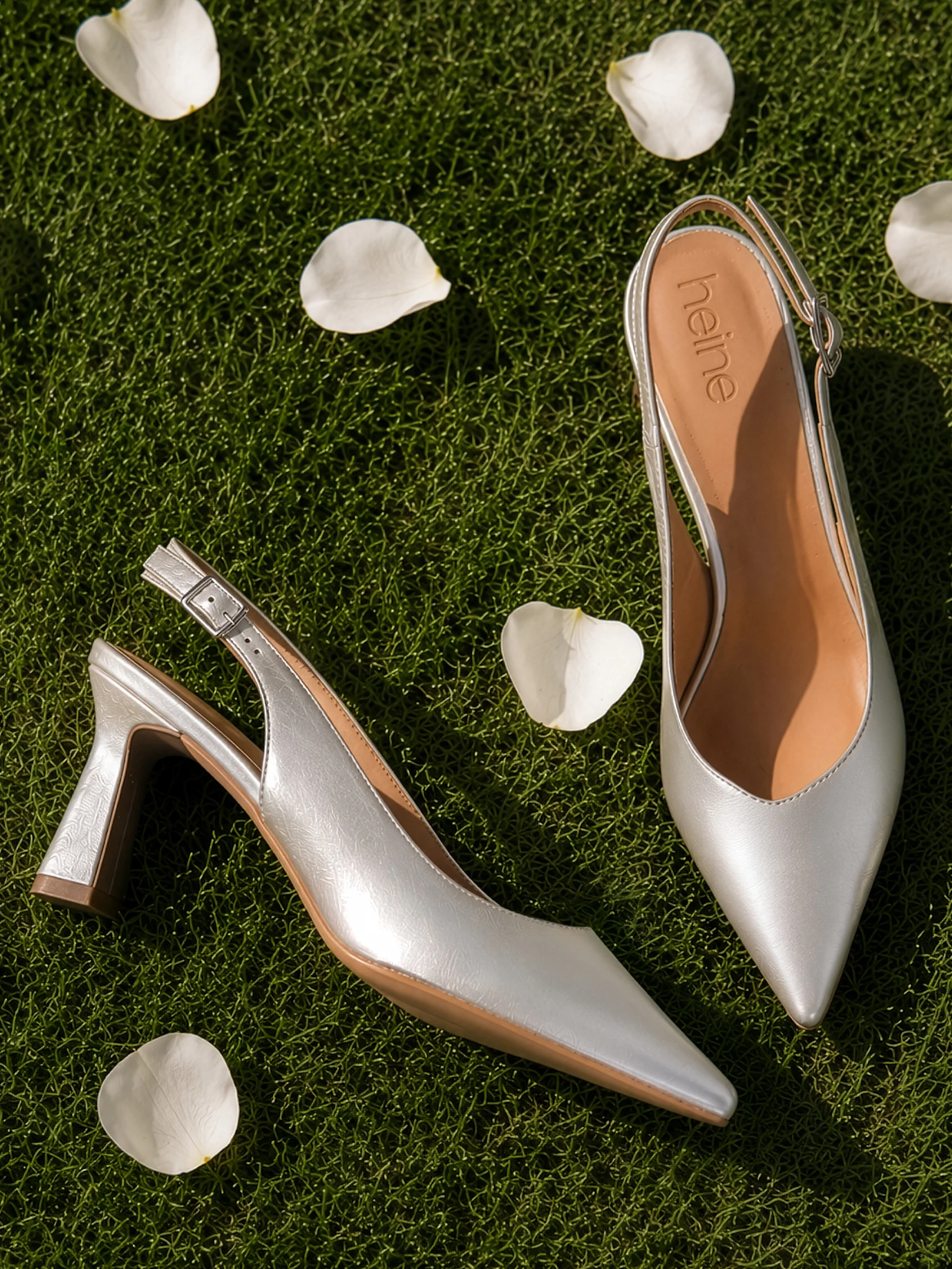 Une paire d'escarpins slingback argentés à talon mi-haut repose sur du gazon vert, entourée de pétales de roses blanches.