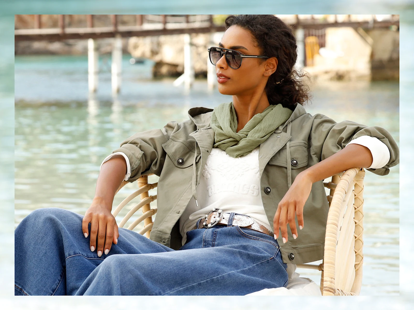 Un mannequin est assis, détendu, dans une chaise en osier au bord de l'eau, vêtu d'une veste vert olive et d'un pull blanc.