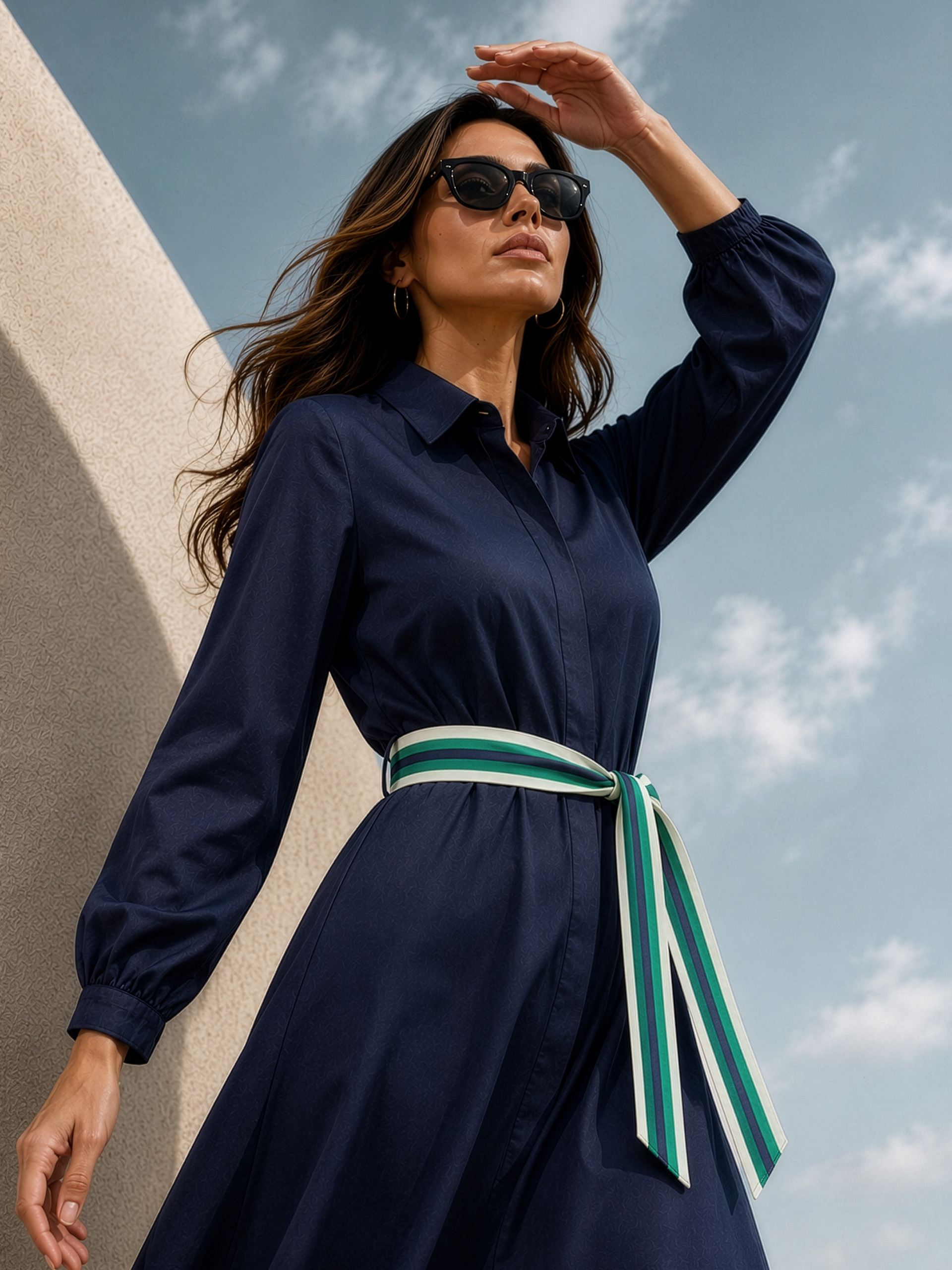 Ein Model steht vor einer hellen Wand, trägt ein dunkelblaues Kleid mit langen Ärmeln und einen gestreiften Gürtel in Grün- und Blautönen. Der Himmel ist leicht bewölkt.
