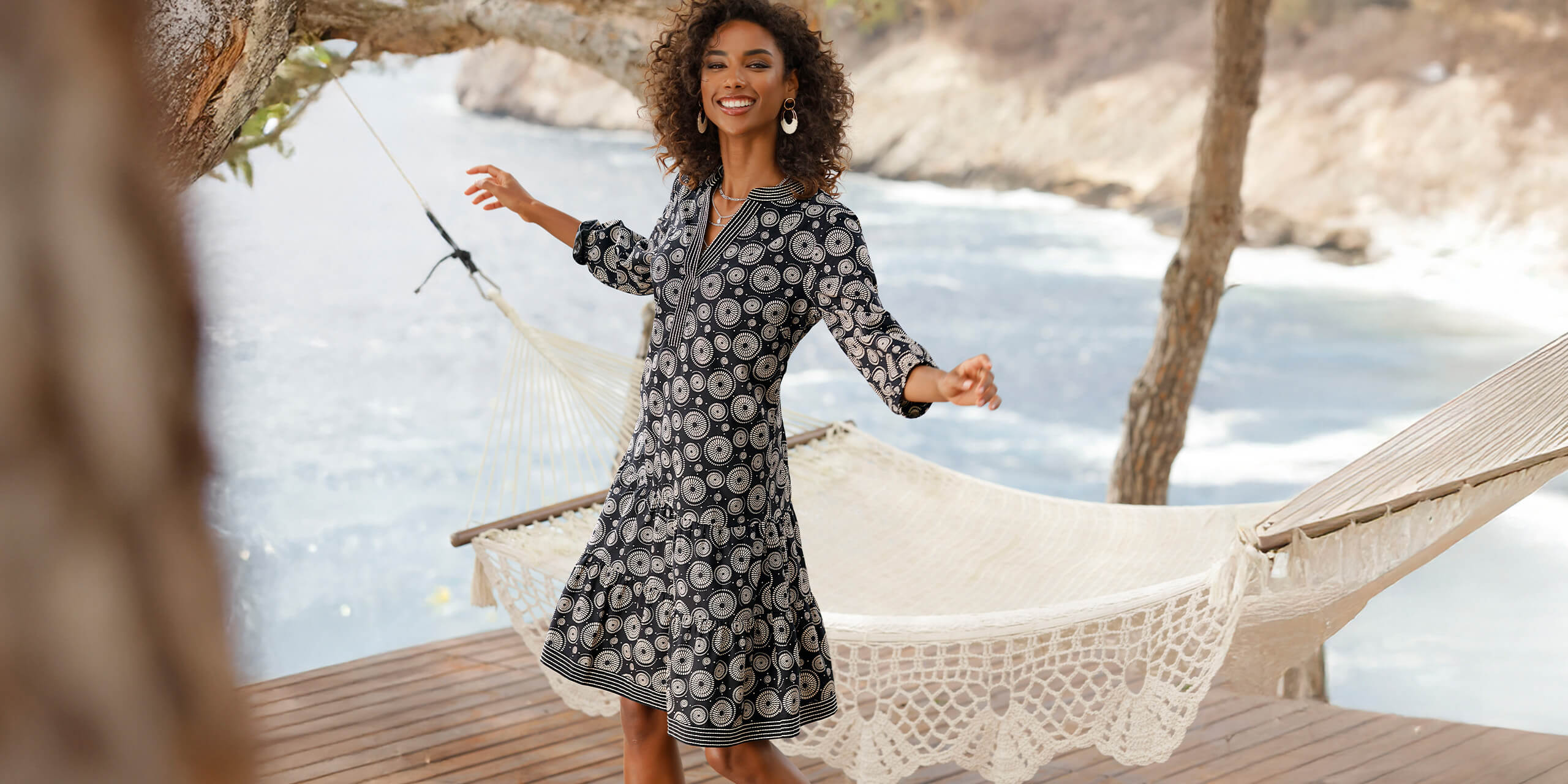 Ein Model trägt ein schwarzes Kleid mit weißem Blumenmuster und steht auf einer Terrasse vor einer Hängematte am Wasser.