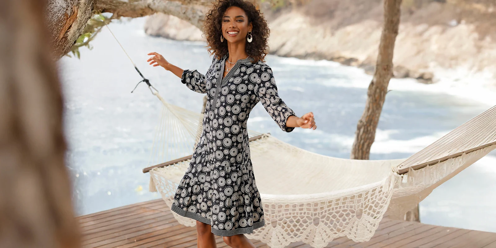 Ein Model trägt ein schwarzes Kleid mit weißem Blumenmuster und steht auf einer Terrasse vor einer Hängematte am Wasser.