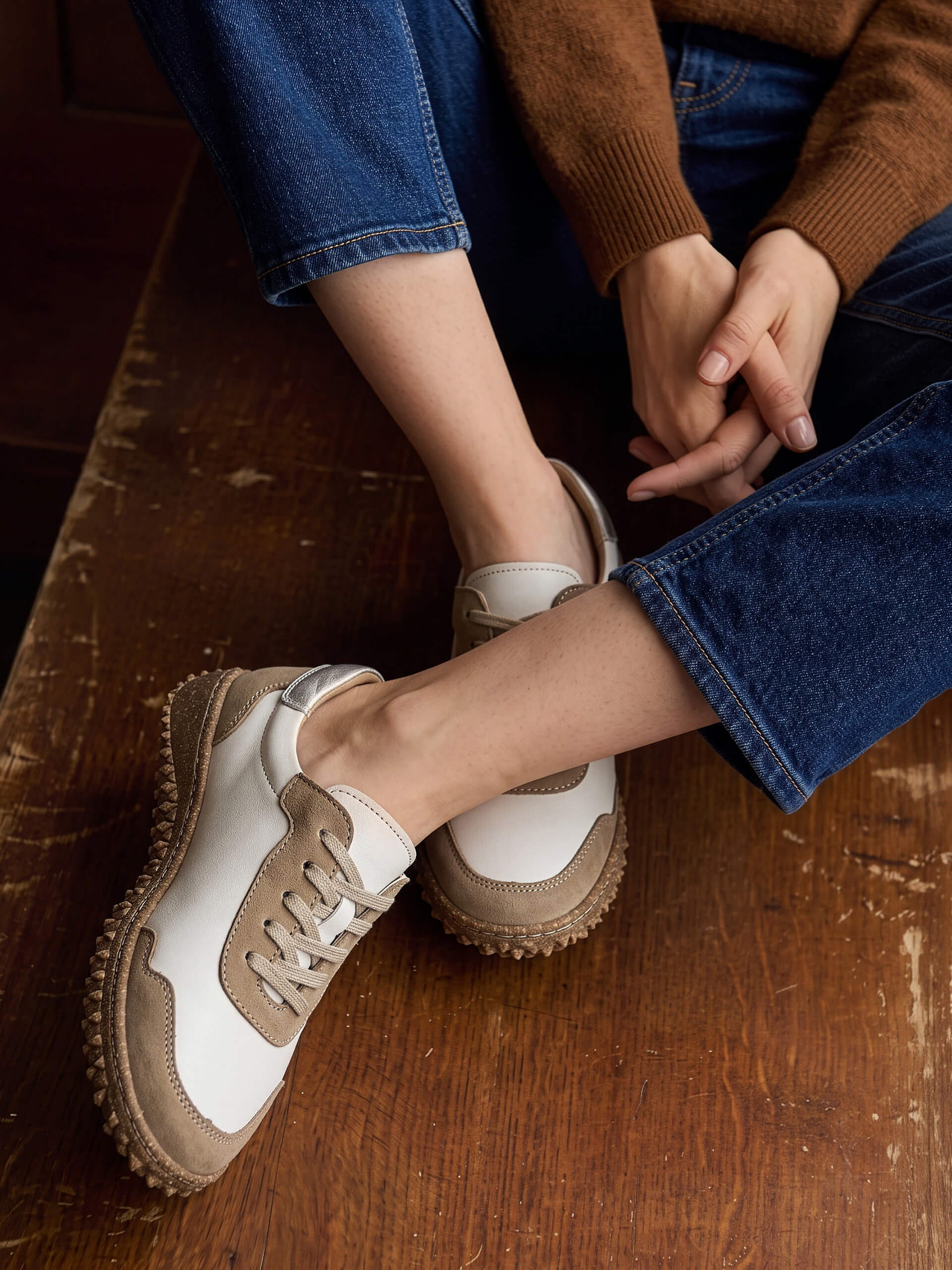 Ein weibliches Model' in blauer Jeans und braunem Pullover trägt weiß-braune Sneaker und sitzt auf einem Holztisch.