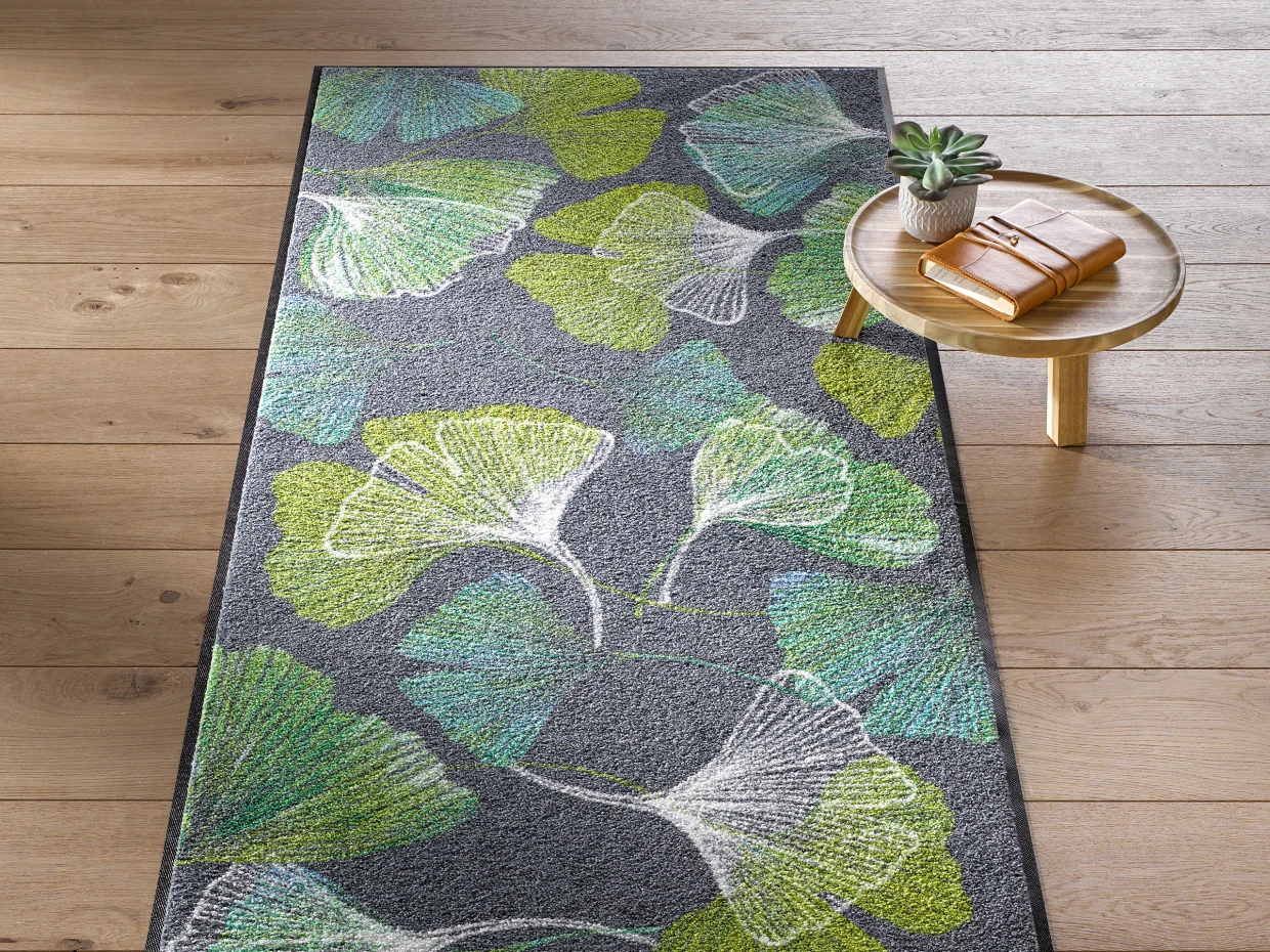 Un paillasson avec un motif de feuilles vertes et grises est posé sur un sol en bois. À côté se trouve une petite table ronde avec une plante et un livre dessus.